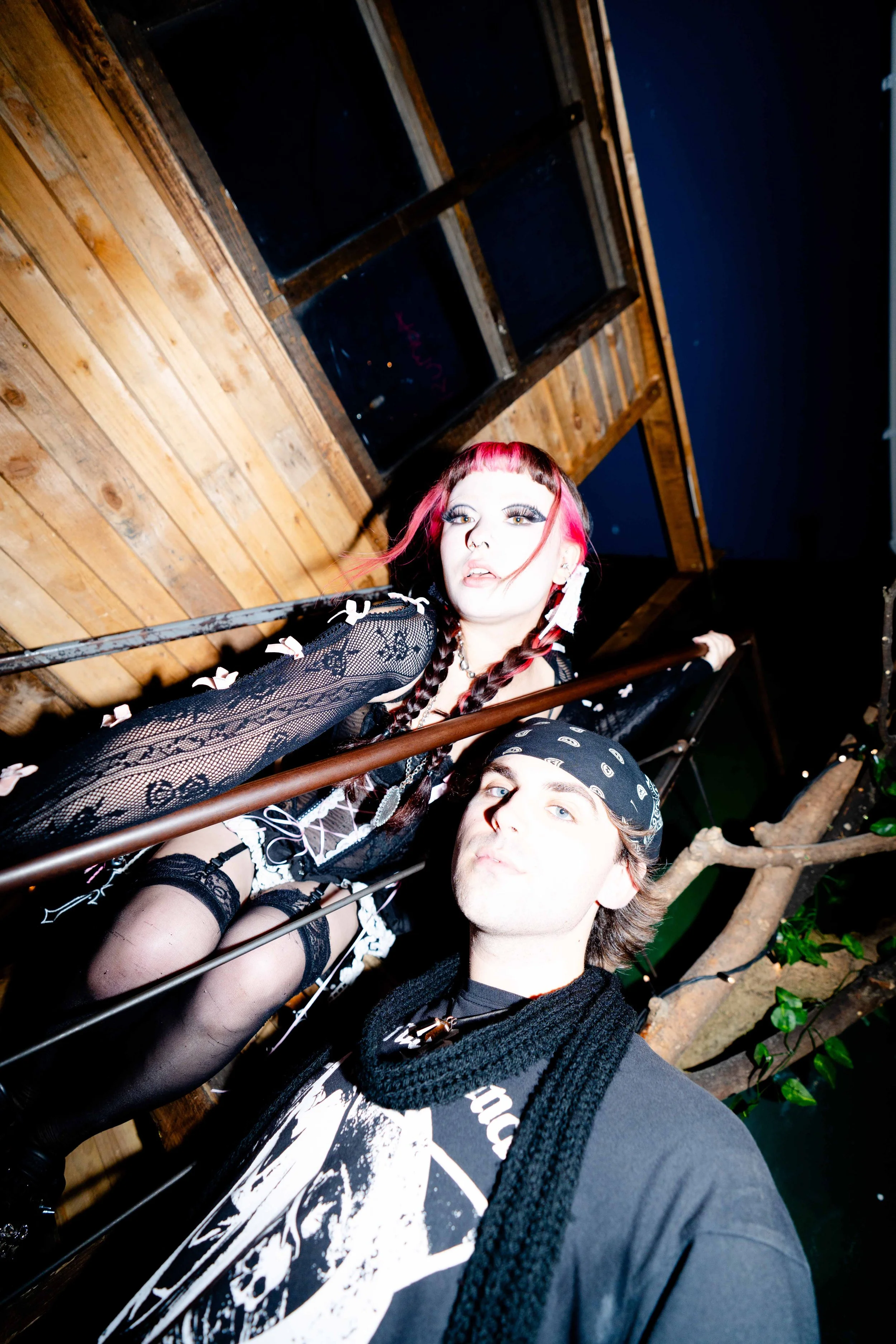 Two people taking a selfie indoors, with a woman featuring pink hair and a man with a bandana, standing on a staircase with wooden walls and a plant in the background.