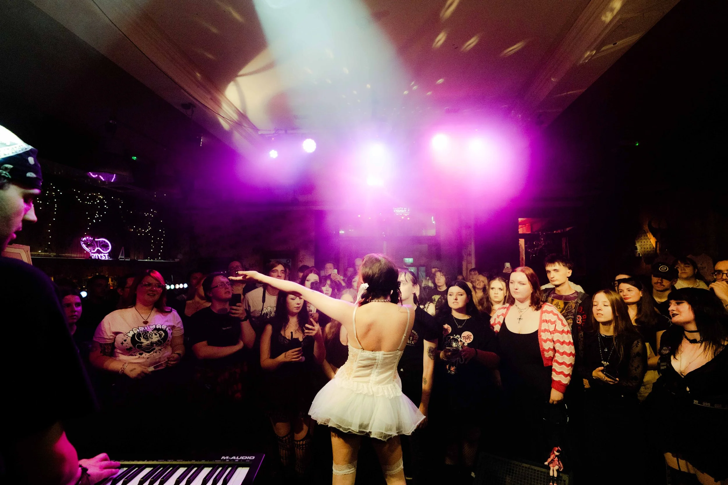 A performer in a white dress and stockings on stage with purple lighting, surrounded by an audience in a dark venue.