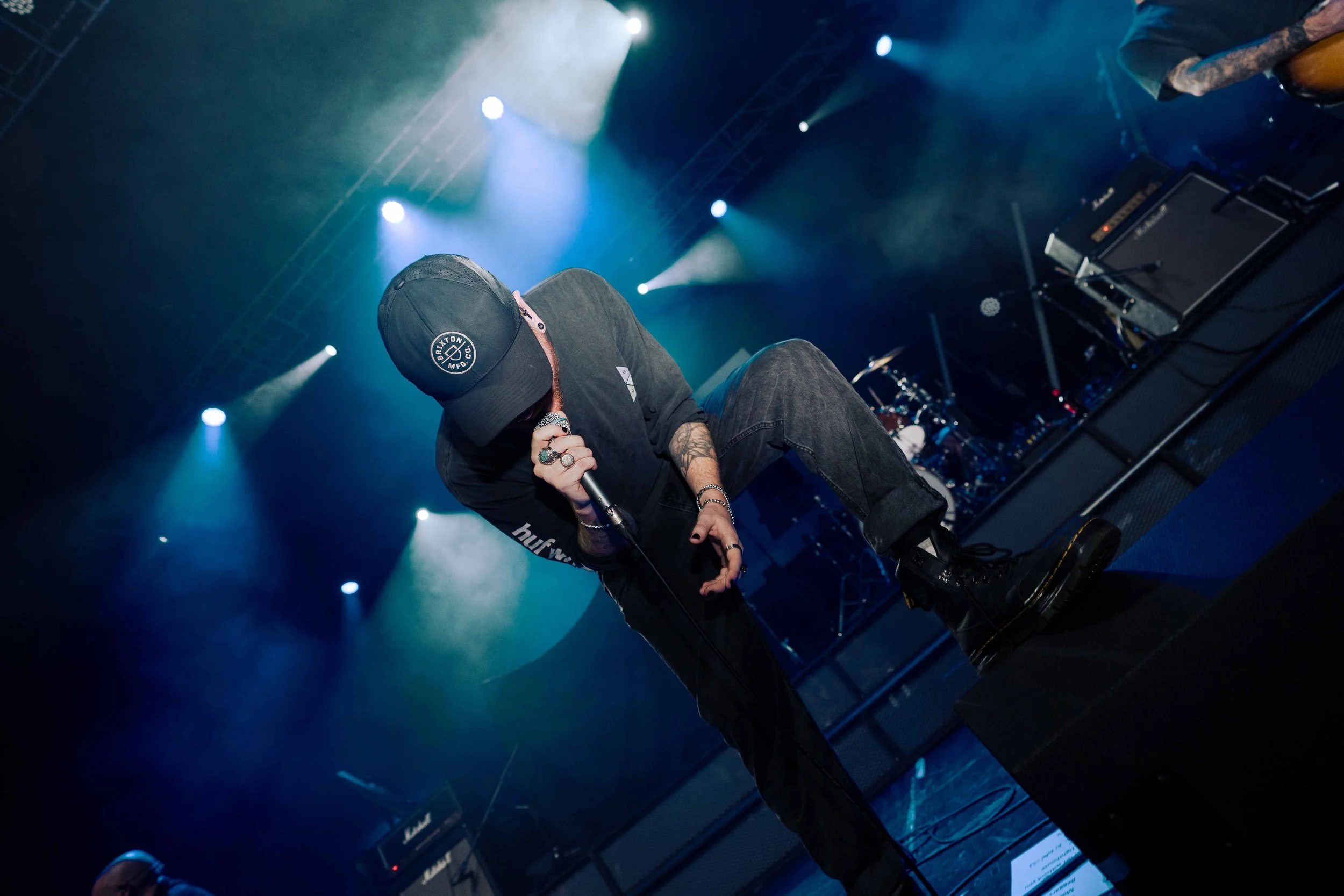 A singer performs on stage wearing a black cap, black clothing, and holding a microphone, with stage lights and musical equipment in the background.