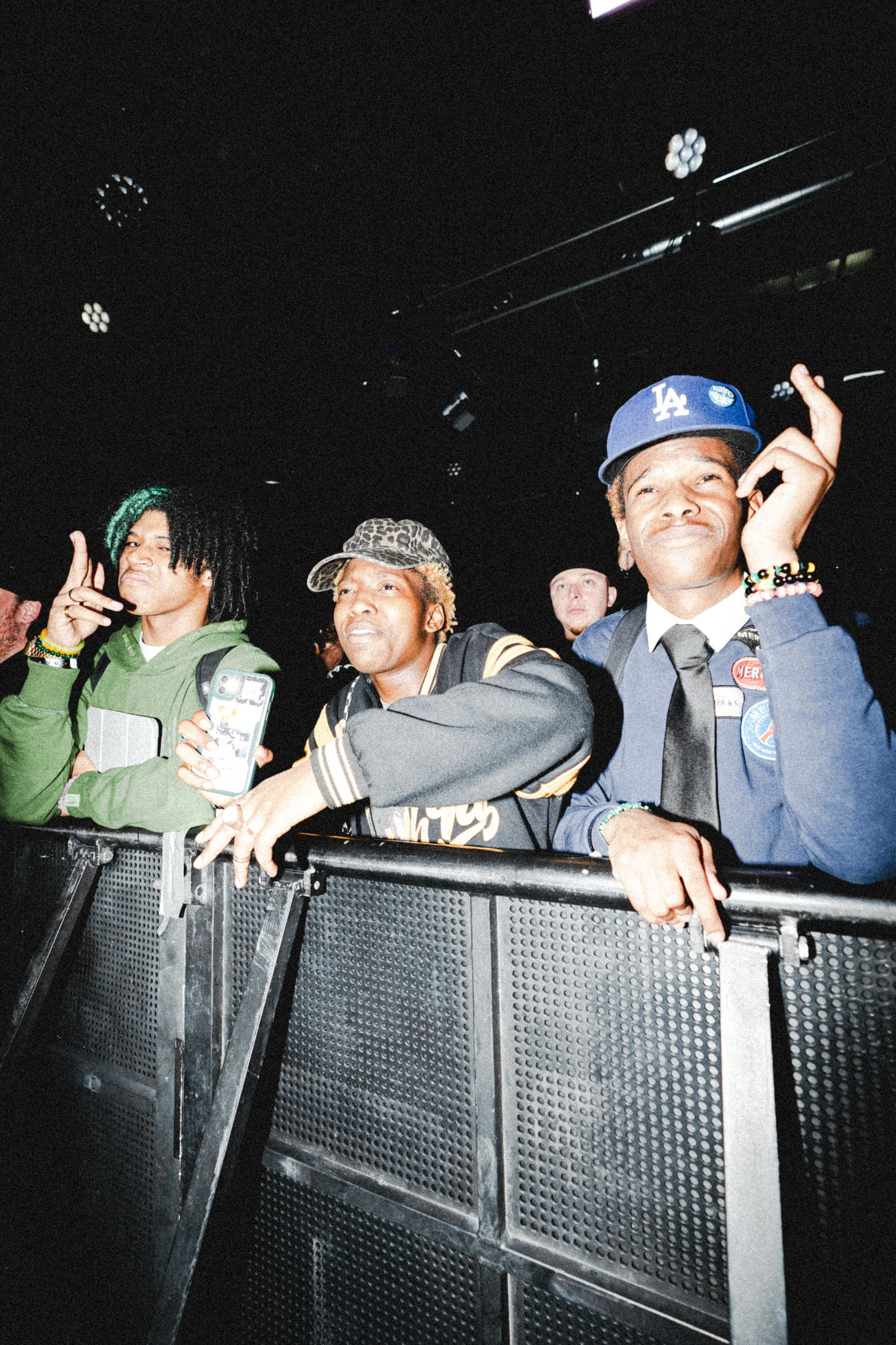 People at a concert or event, standing behind a barrier, with some raising their hands or making gestures.