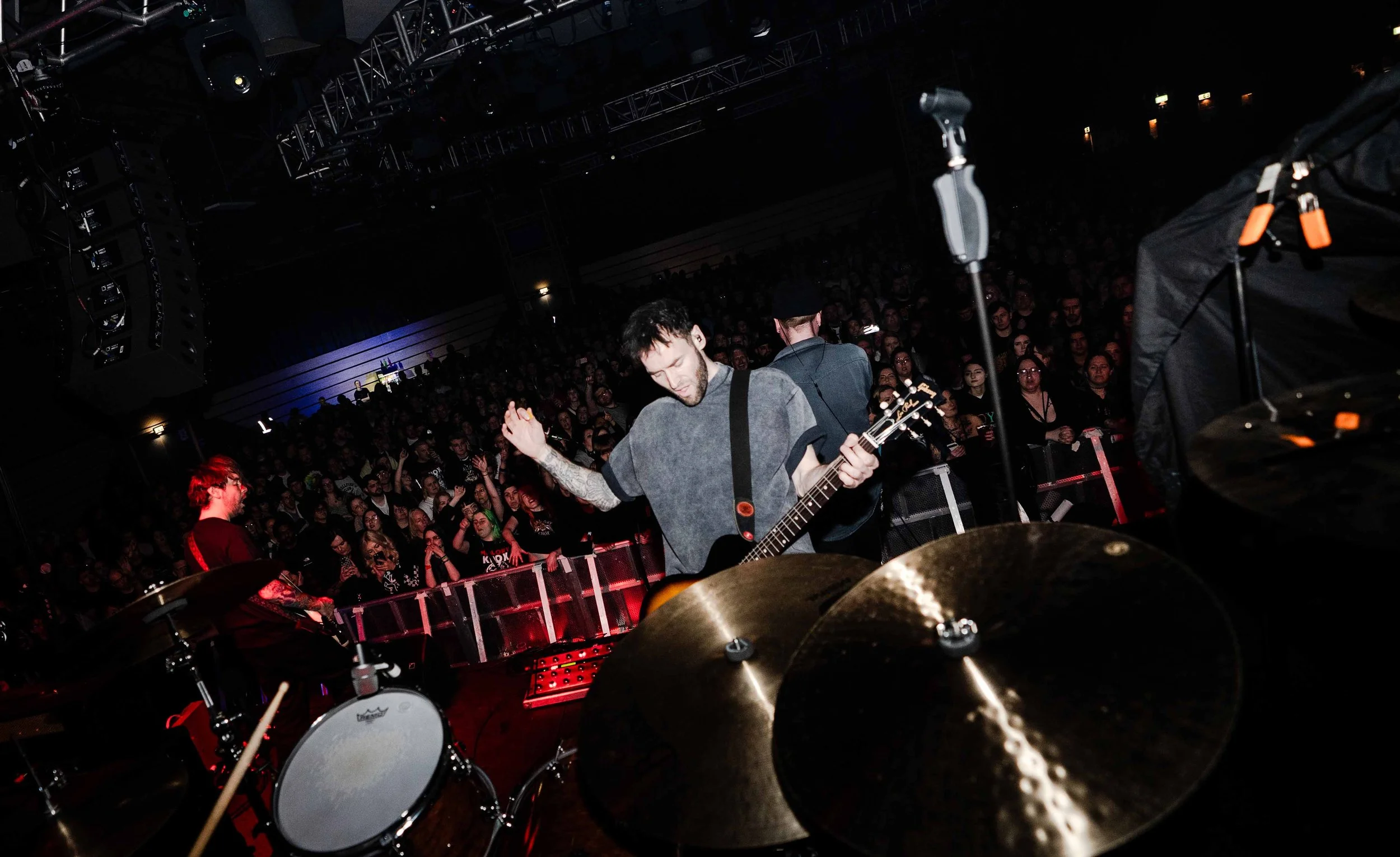A band performing on stage at a concert, with a crowd watching in the background. The image is taken from behind the drum set showing a guitarist and a drummer.