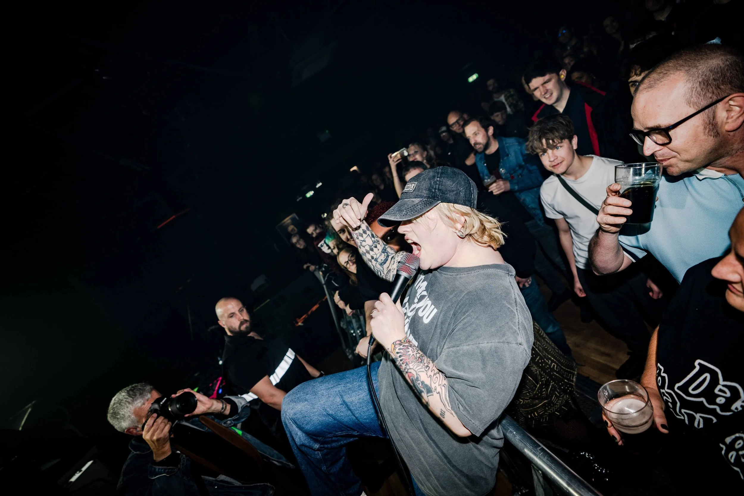 A woman wearing a baseball cap and oversized t-shirt singing into a microphone at a concert, surrounded by an audience enjoying the performance.