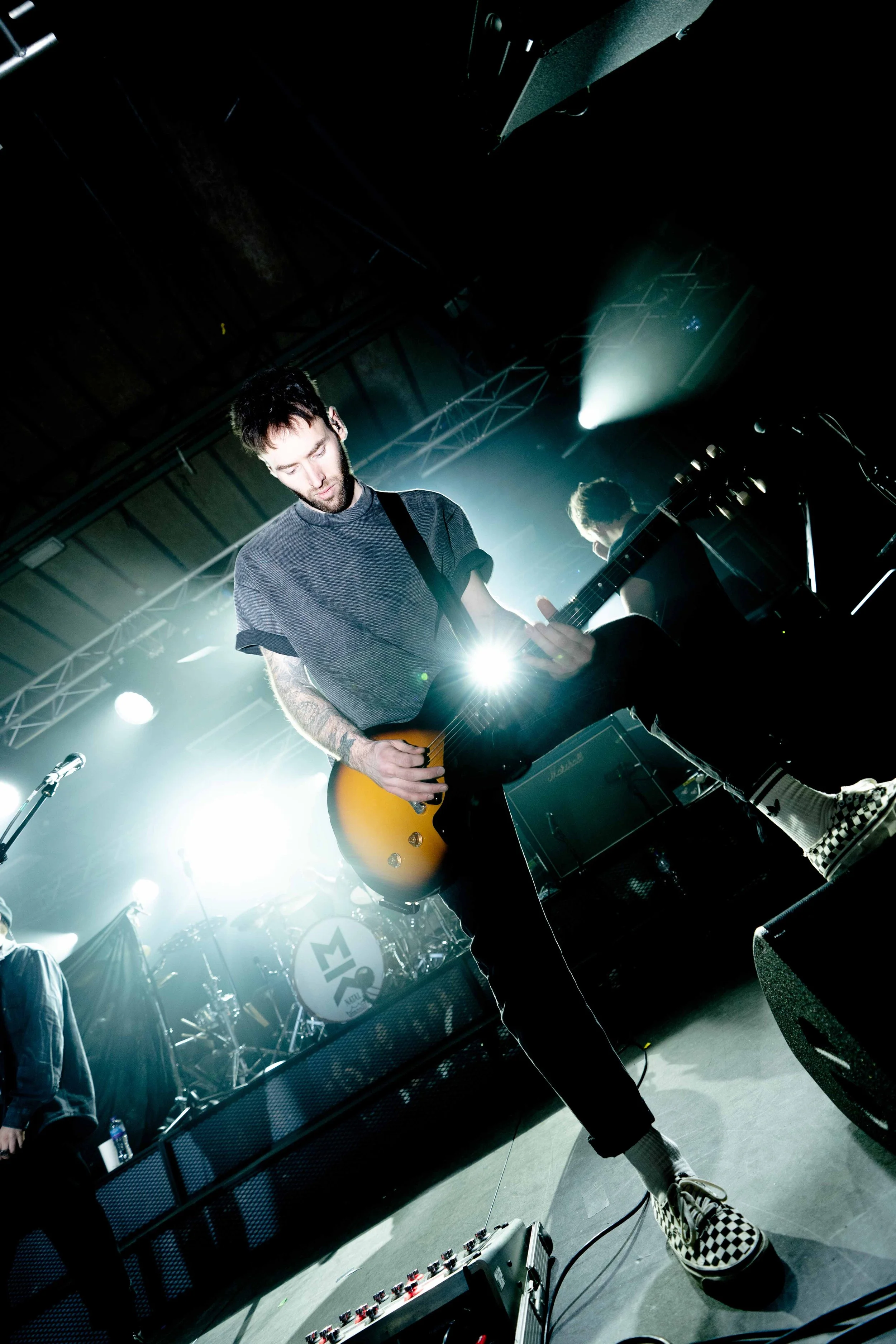 A musician playing an electric guitar on stage during a concert, with band equipment and bright stage lights in the background.