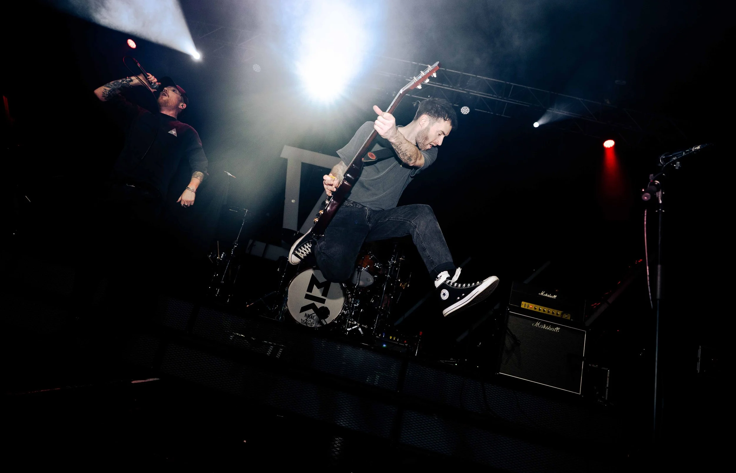 A musician jumps in the air while playing an electric guitar on stage during a concert, with another performer singing into a microphone in the background and stage lights above.