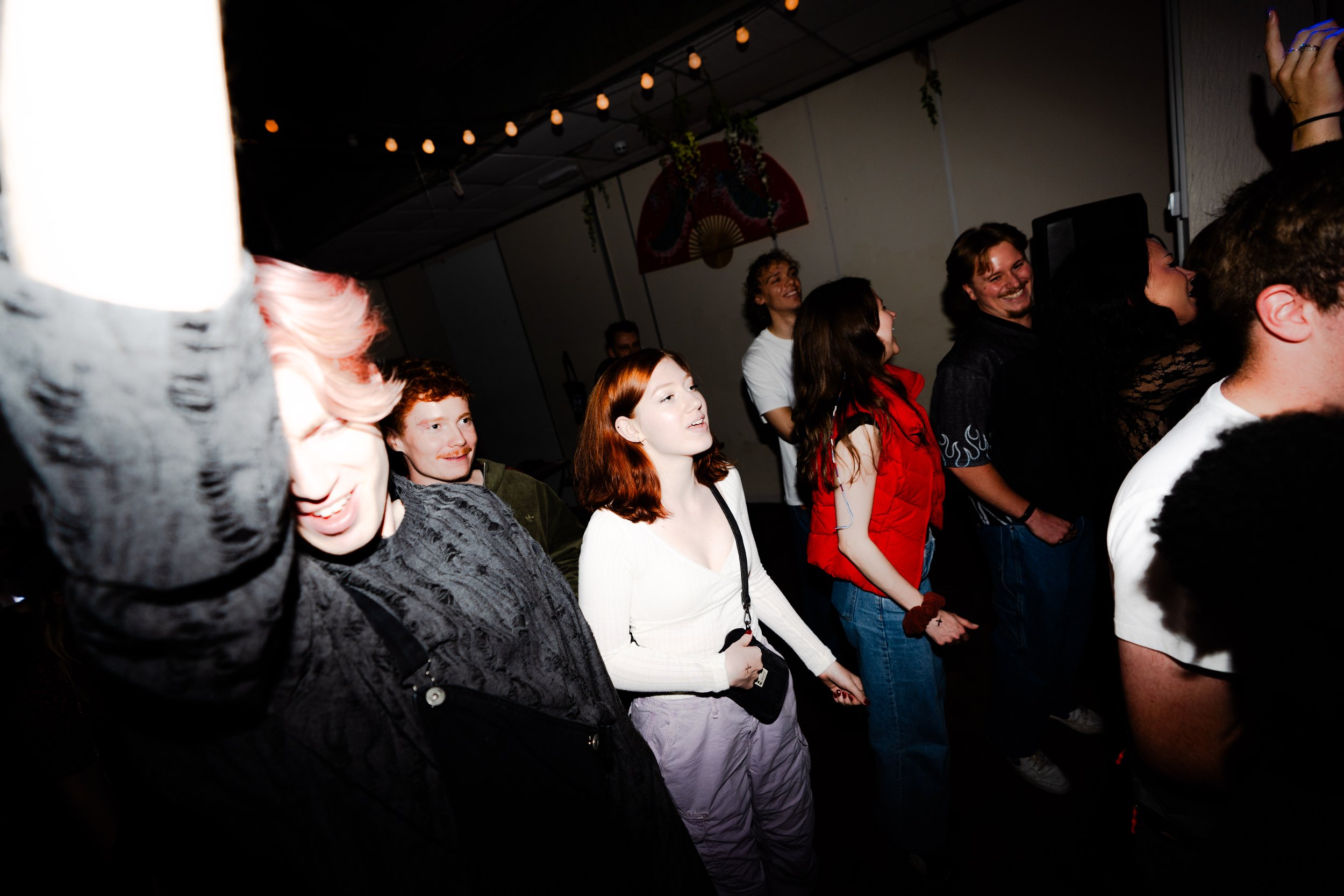Group of young adults enjoying a concert or event, some smiling and laughing, some singing along, under string lights and decorative hangings.