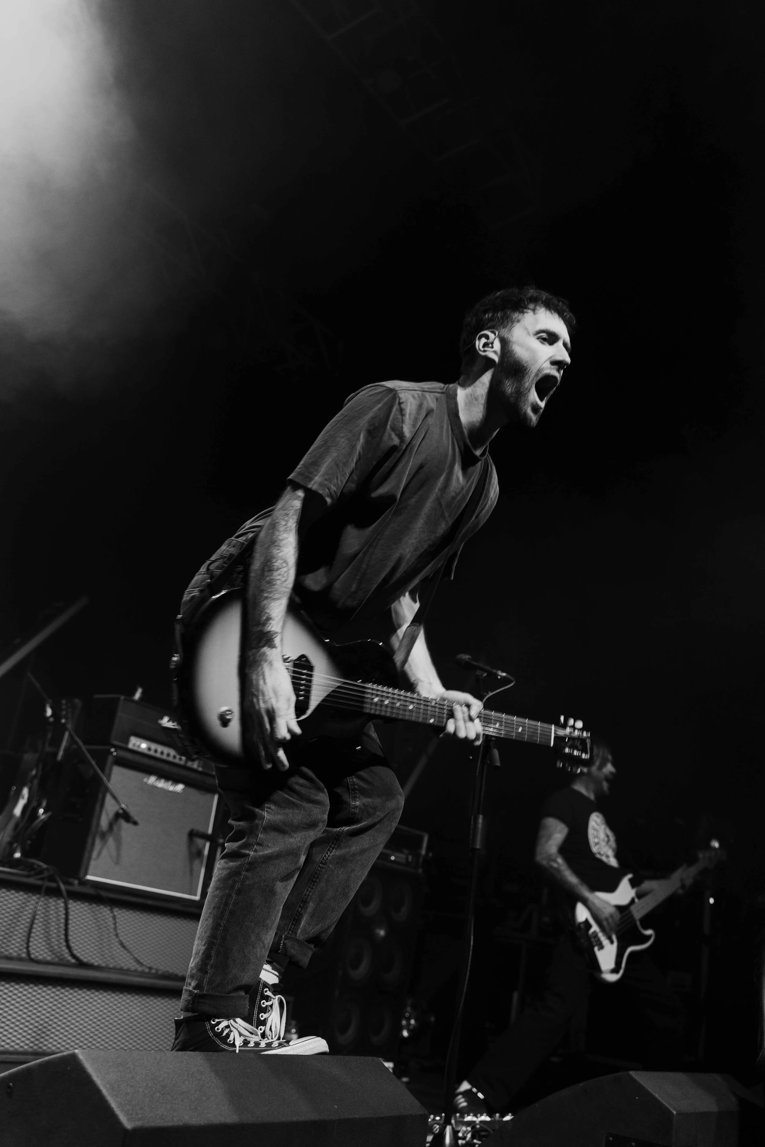A black and white photo of a man playing an electric guitar on stage, mid-performance, with his mouth open as if singing or shouting.