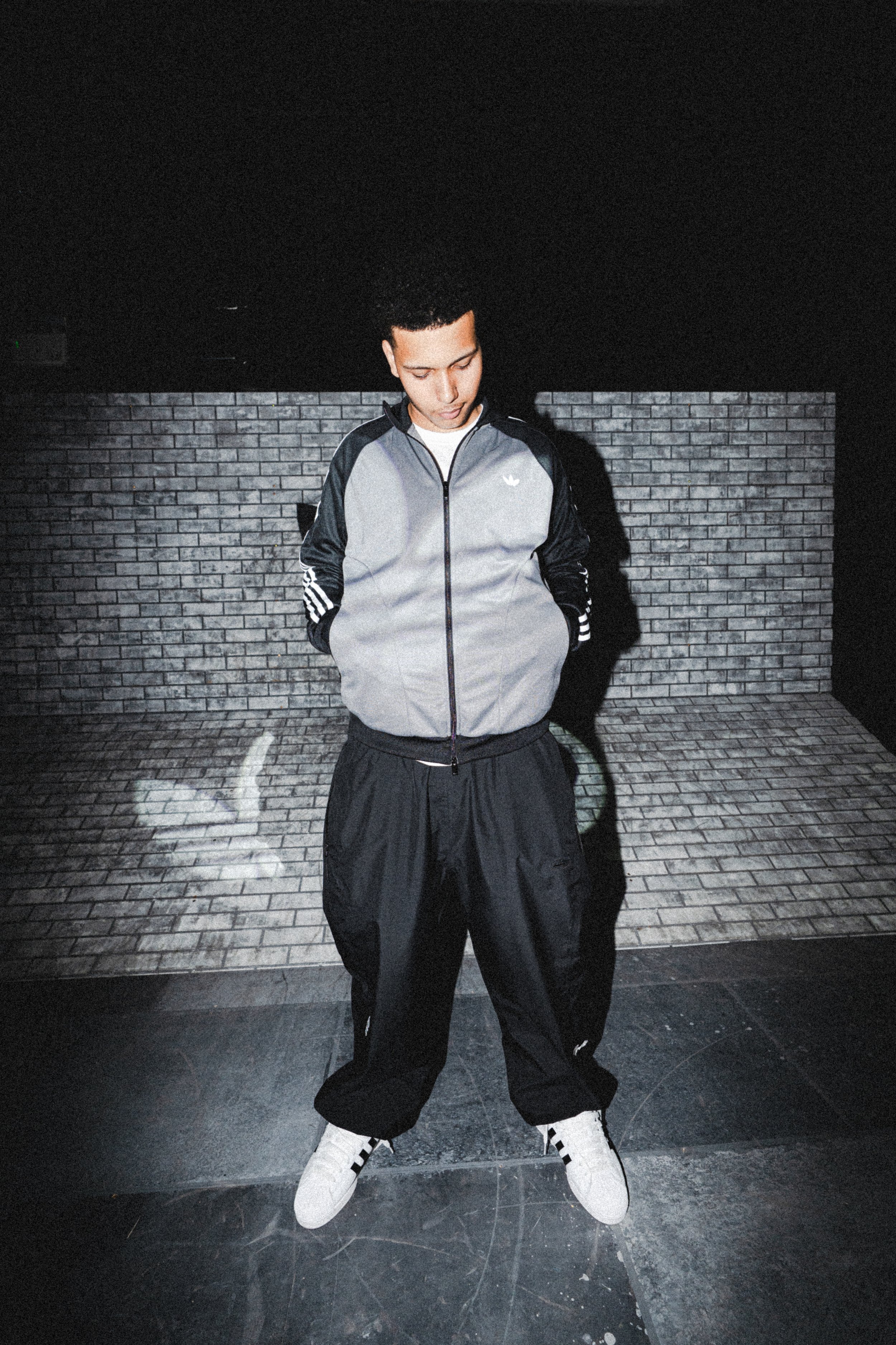 A young man standing against a brick wall at night, dressed in a gray and black Adidas jacket, black pants, and white sneakers, looking down.