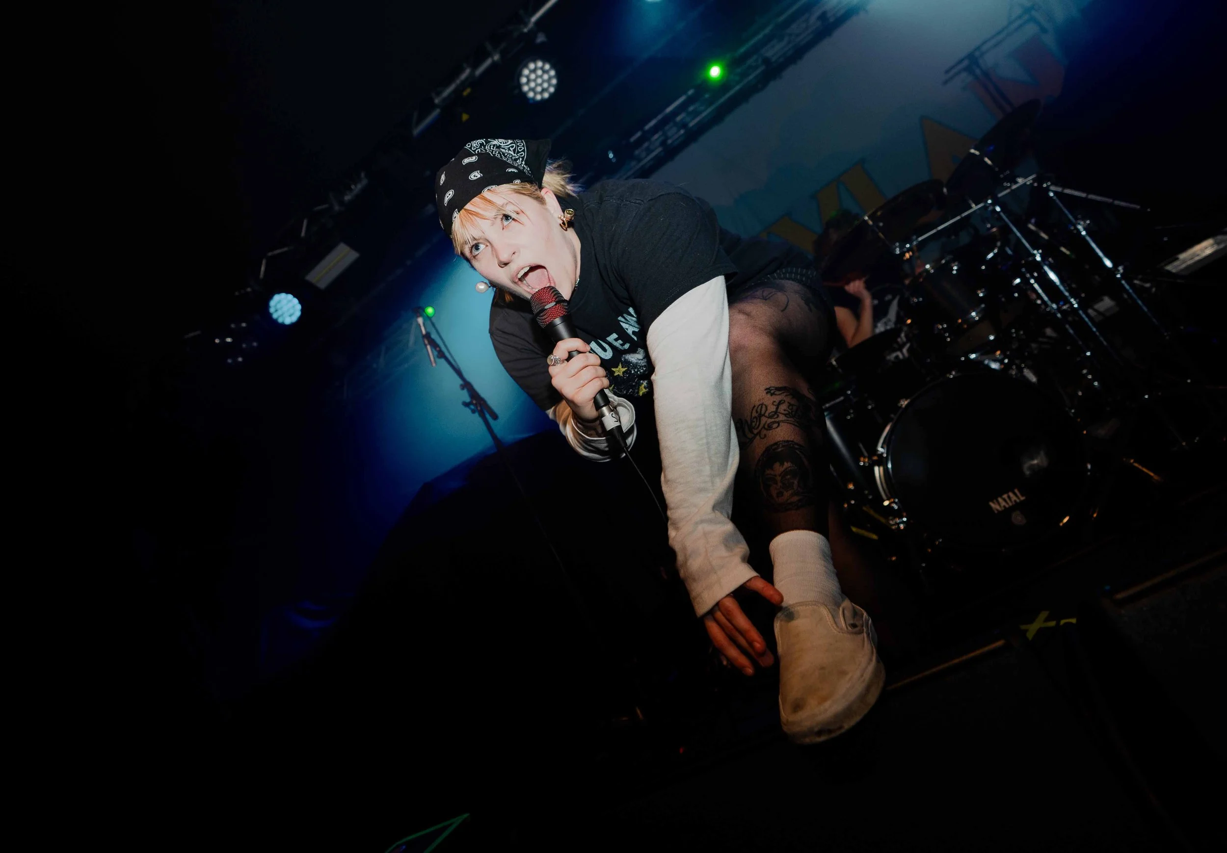 A female singer with short blonde hair, wearing a black bandana, black and white clothing, and patterned tights, crouches on a stage while holding a microphone, performing passionately with stage lights and musical equipment in the background.