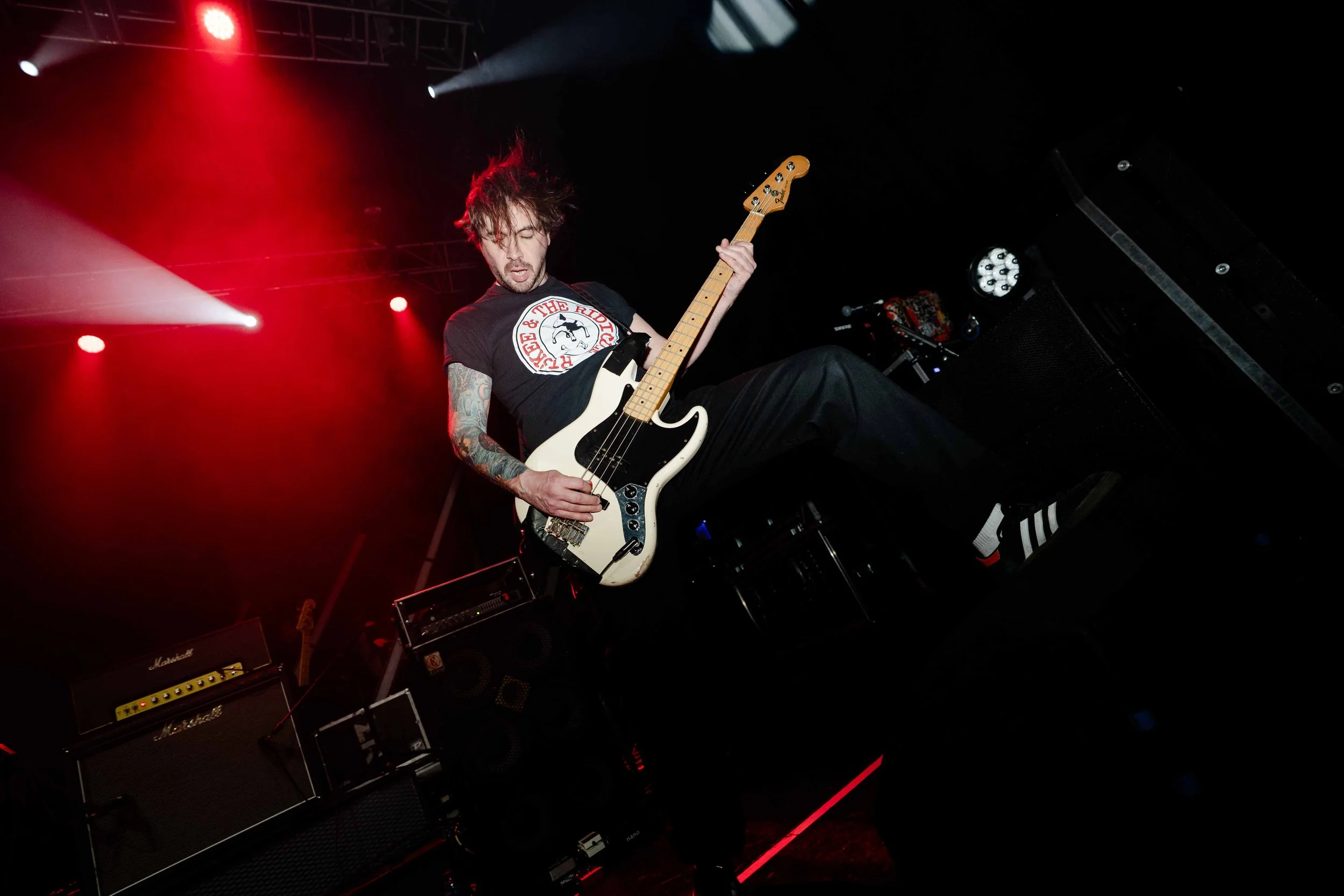 Musician playing electric bass guitar on stage with red and white stage lighting, wearing a black t-shirt and black pants.