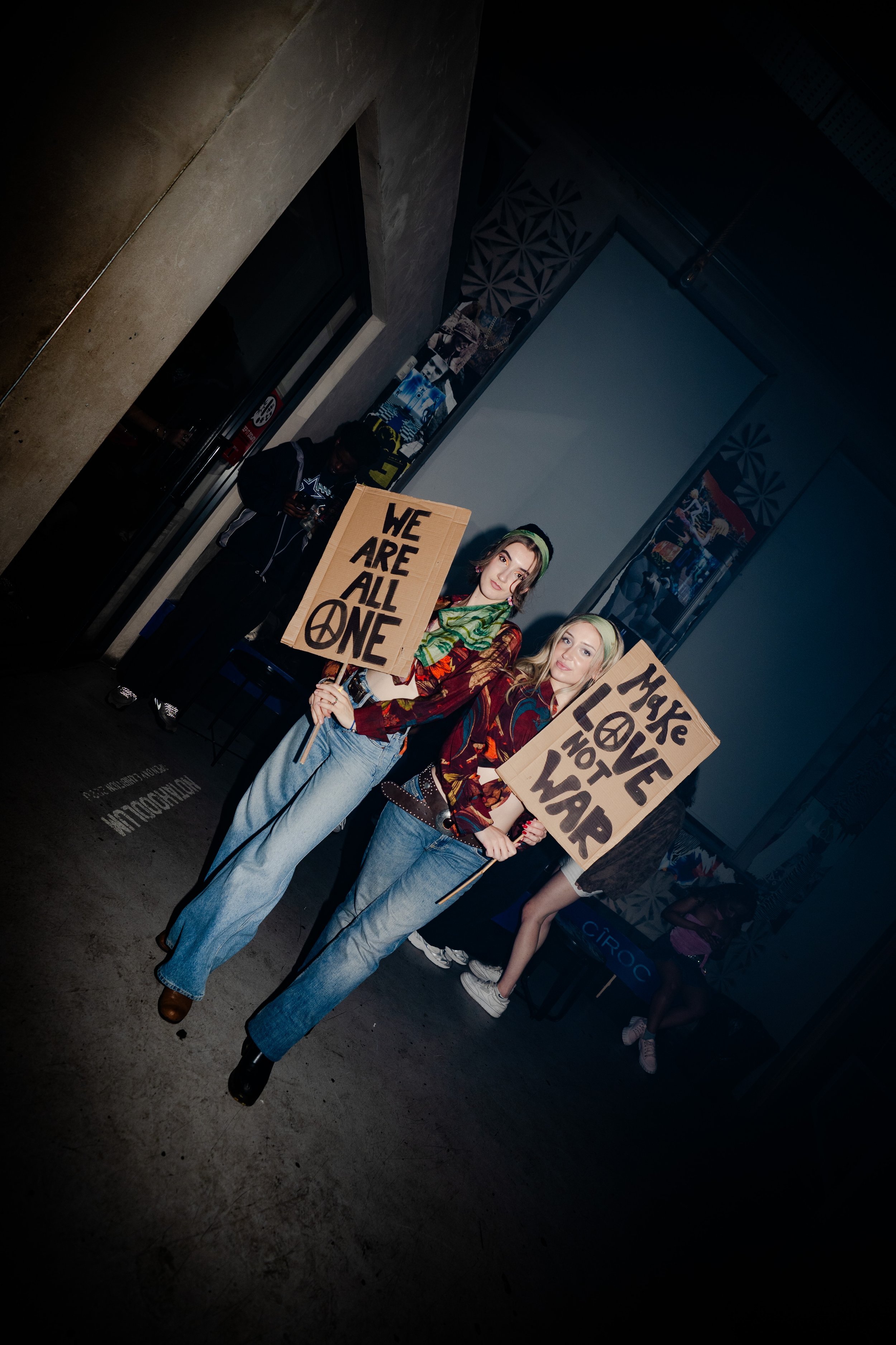Two women protesting with cardboard signs, one reading 'We Are All One' and the other 'Make Love Not War,' standing indoors with other people in a dimly lit setting.