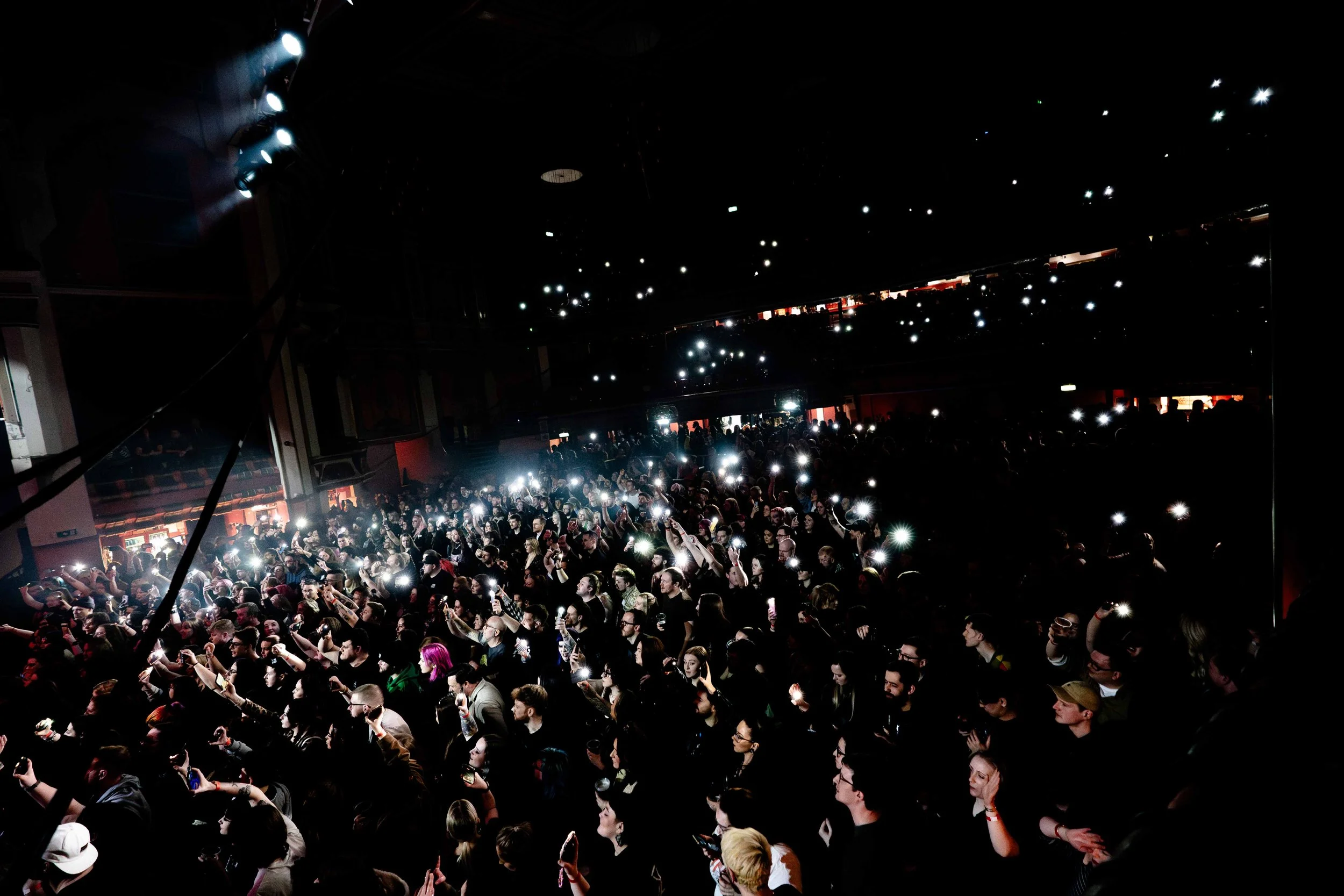 Crowd of people at a concert or event, holding up phones with flashlights on in a dark venue.