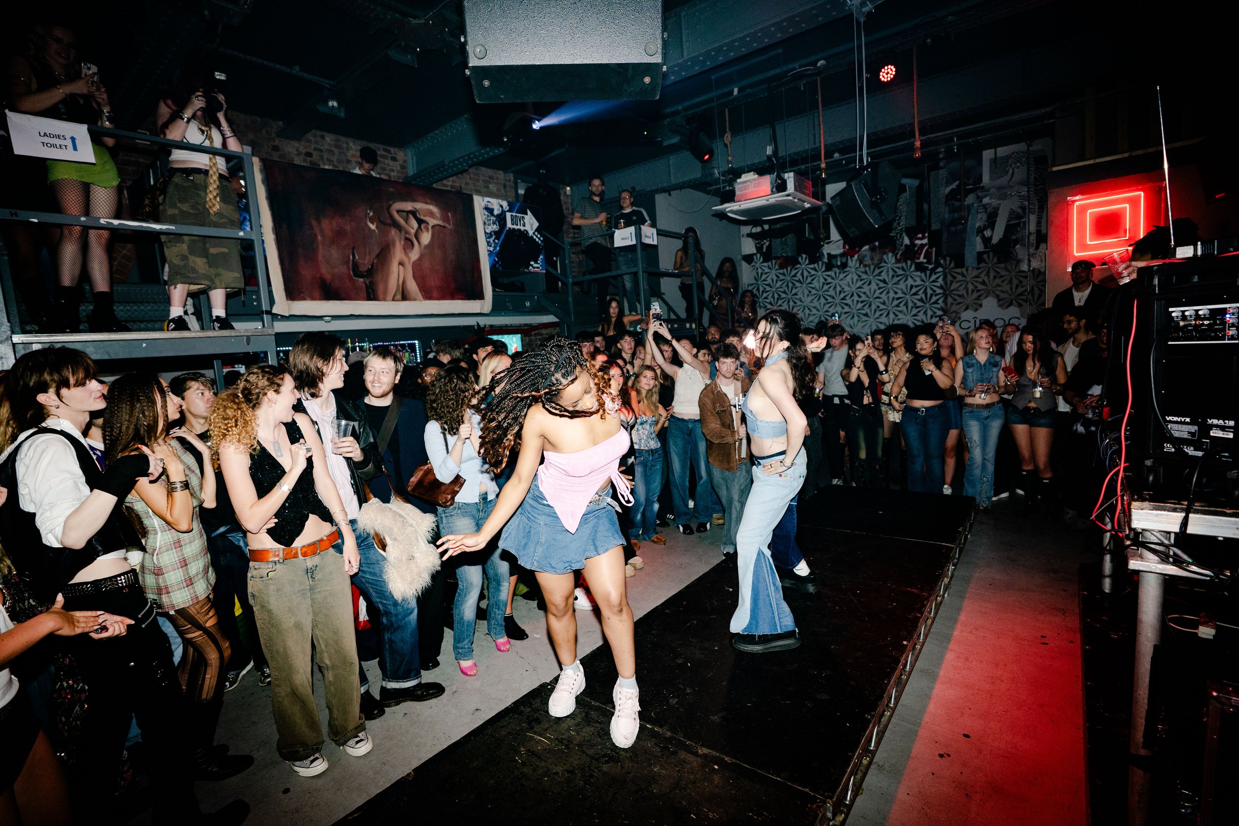 People dancing on a small stage in a crowded nightclub with a DJ booth, artwork, and neon sign in the background.