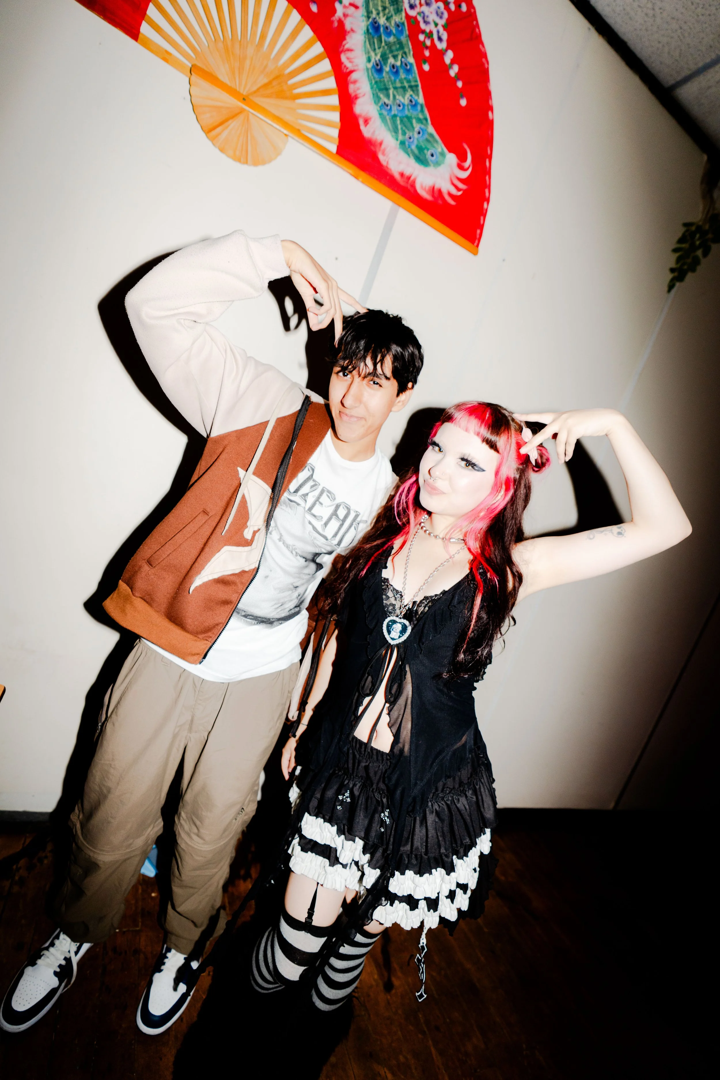 young man and woman posing together indoors, the woman with pink and black hair wearing a black Gothic-style dress with striped stockings, the man wearing casual beige pants, a graphic t-shirt, and a brown and beige jacket, with a colorful Asian-styl