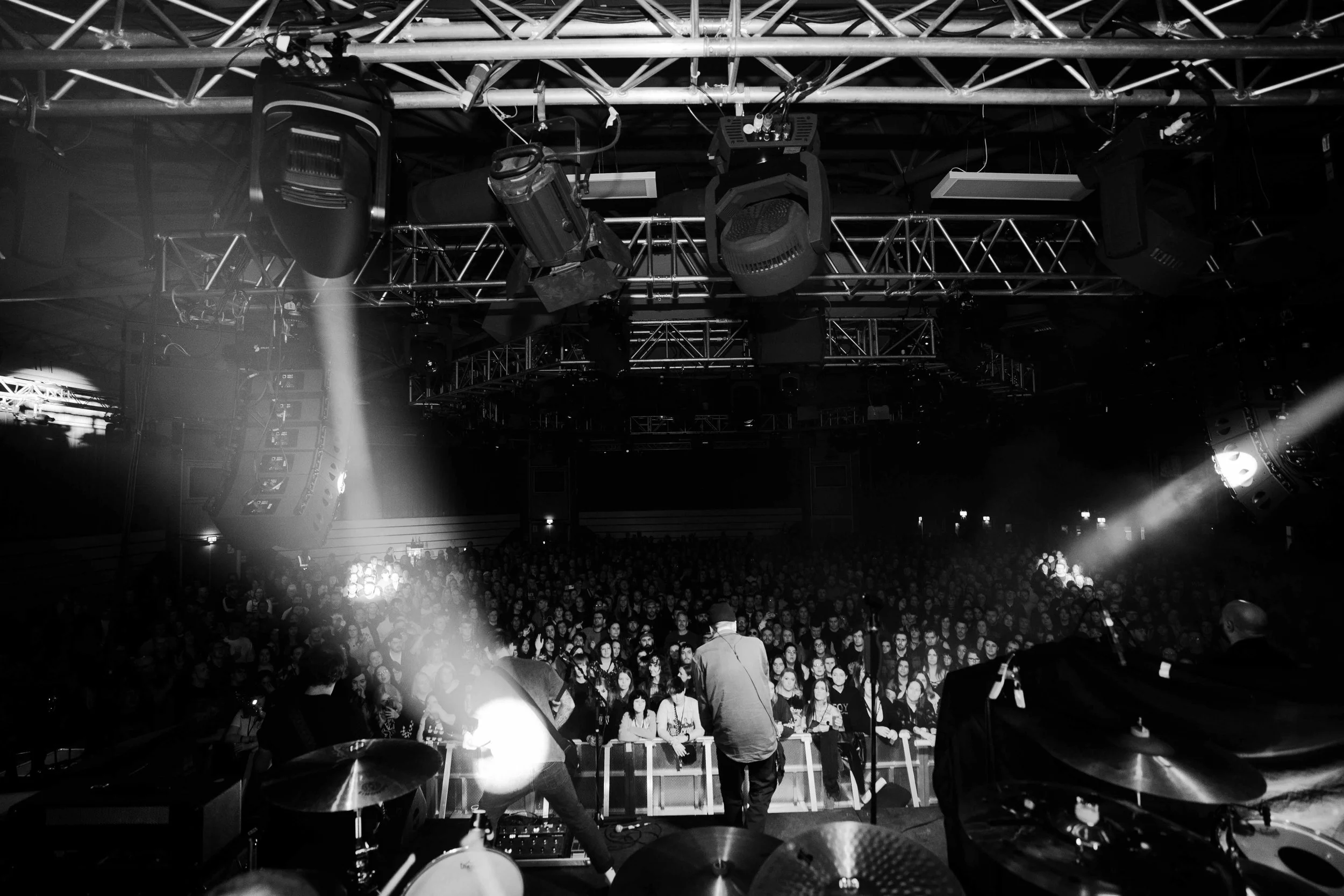 Black and white photo of a live music concert from the stage with a DJ or performers. The crowd is densely packed, facing the stage in a dark indoor venue with stage lighting, speakers, and equipment overhead.