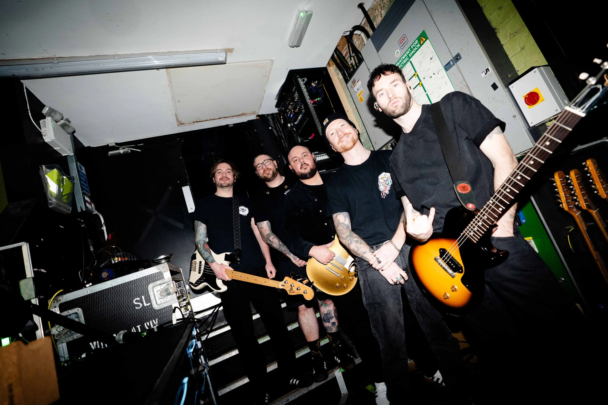 Five men in black and dark clothing, some with tattoos, standing in a backstage or rehearsal room holding guitars, with equipment and cables around.