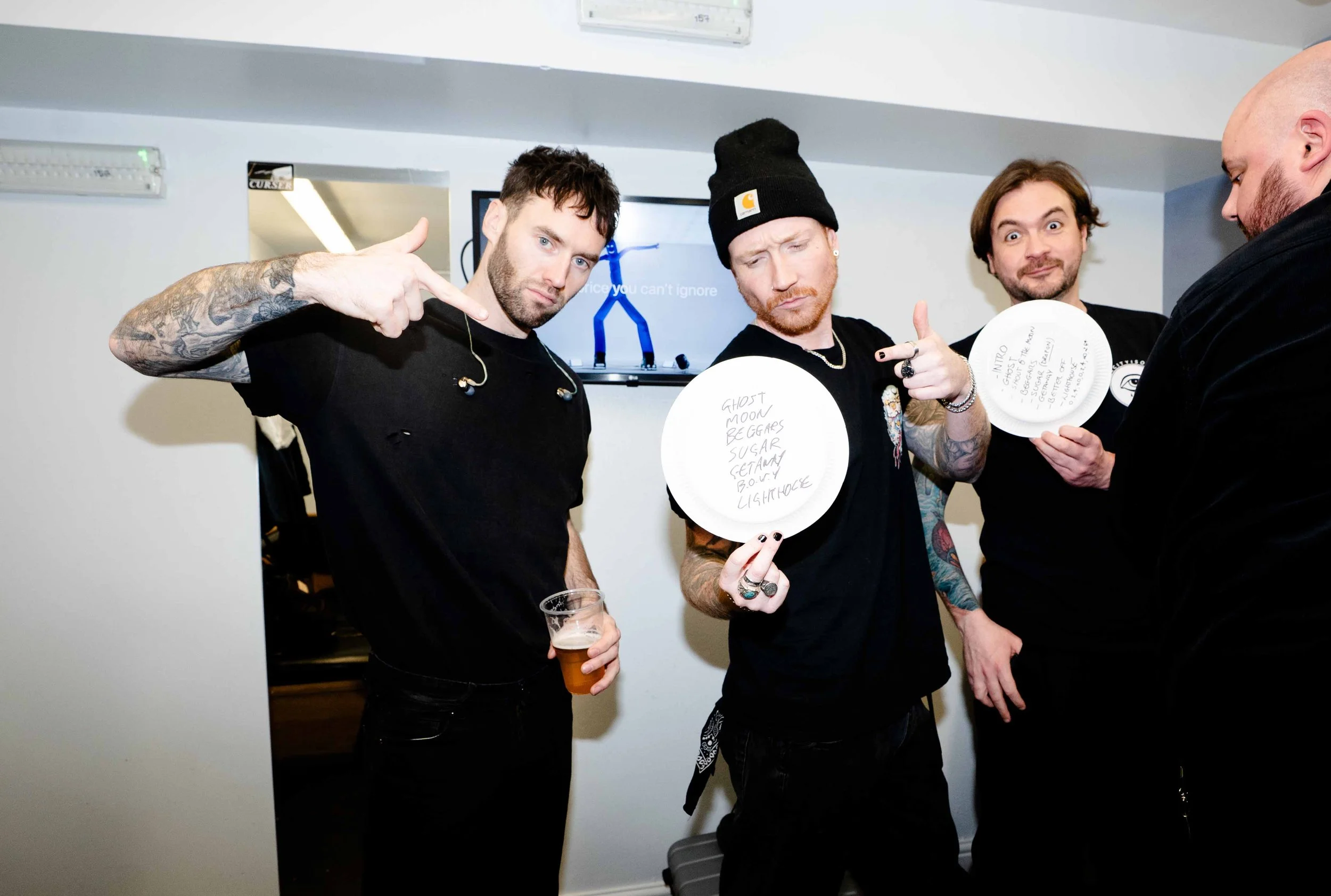 Four men with tattoos and dark clothing standing together, holding paper plates with handwritten song titles, one is drinking a beer, and one is showing a hand gesture; they appear to be at a casual gathering or event.