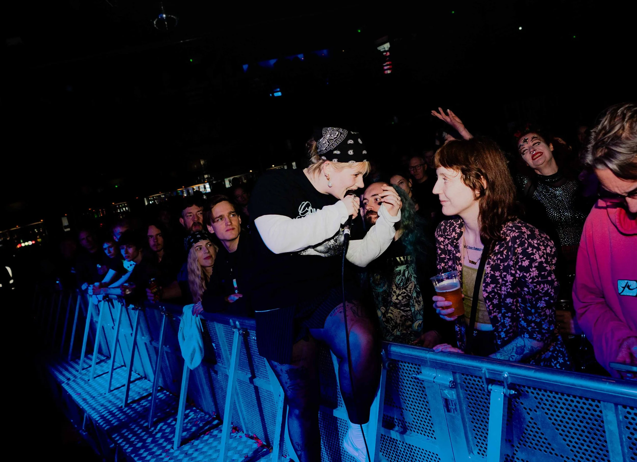 A woman with blonde hair, wearing a bandana and black t-shirt, singing into a microphone at a crowded concert or music event. She is leaning over a metal barrier to interact with audience members.