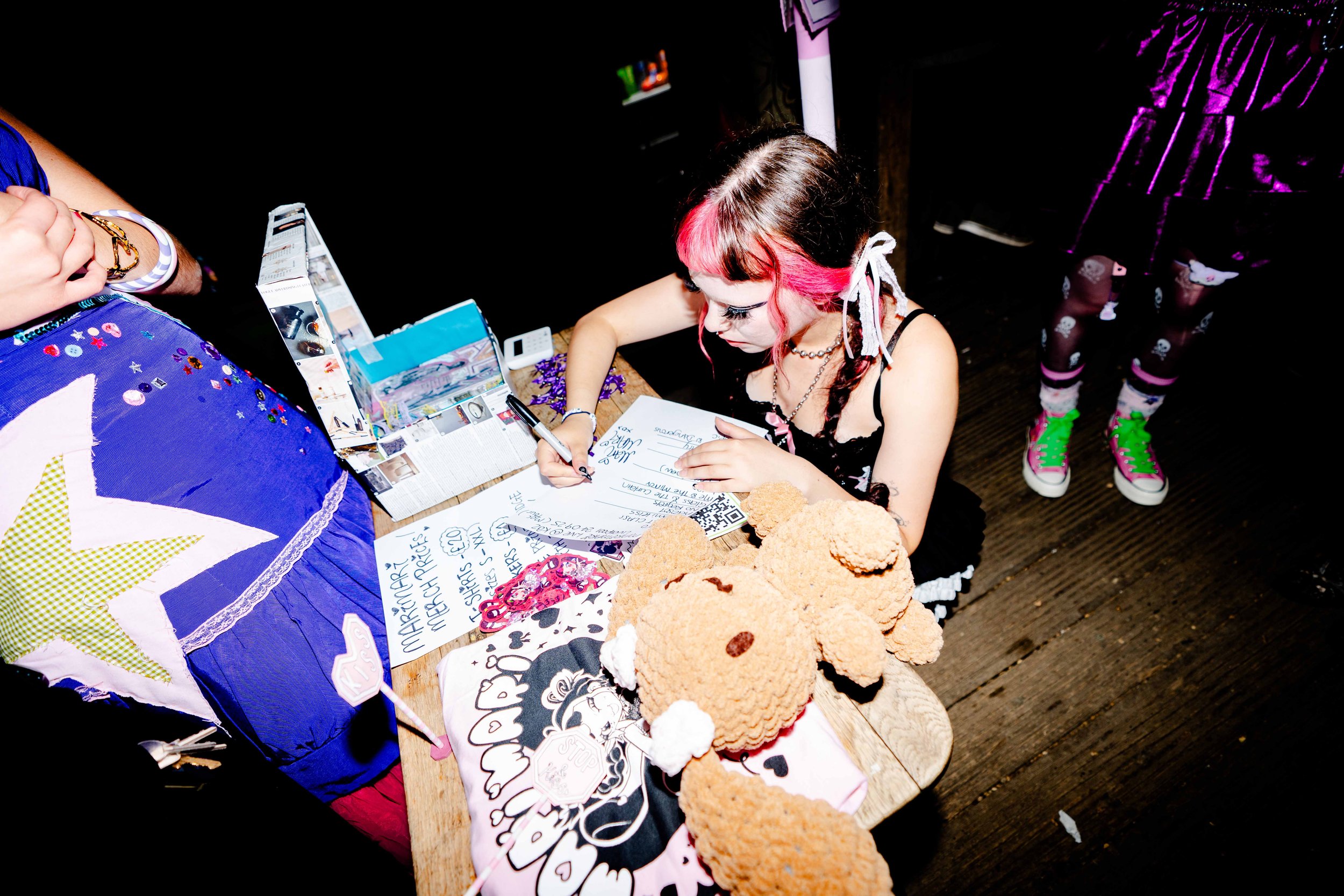 A young woman with pink and black hair signing autographs at a table covered with colorful merchandise, plush toys, and signs, with a girl in a purple dress and neon green shoes standing nearby.