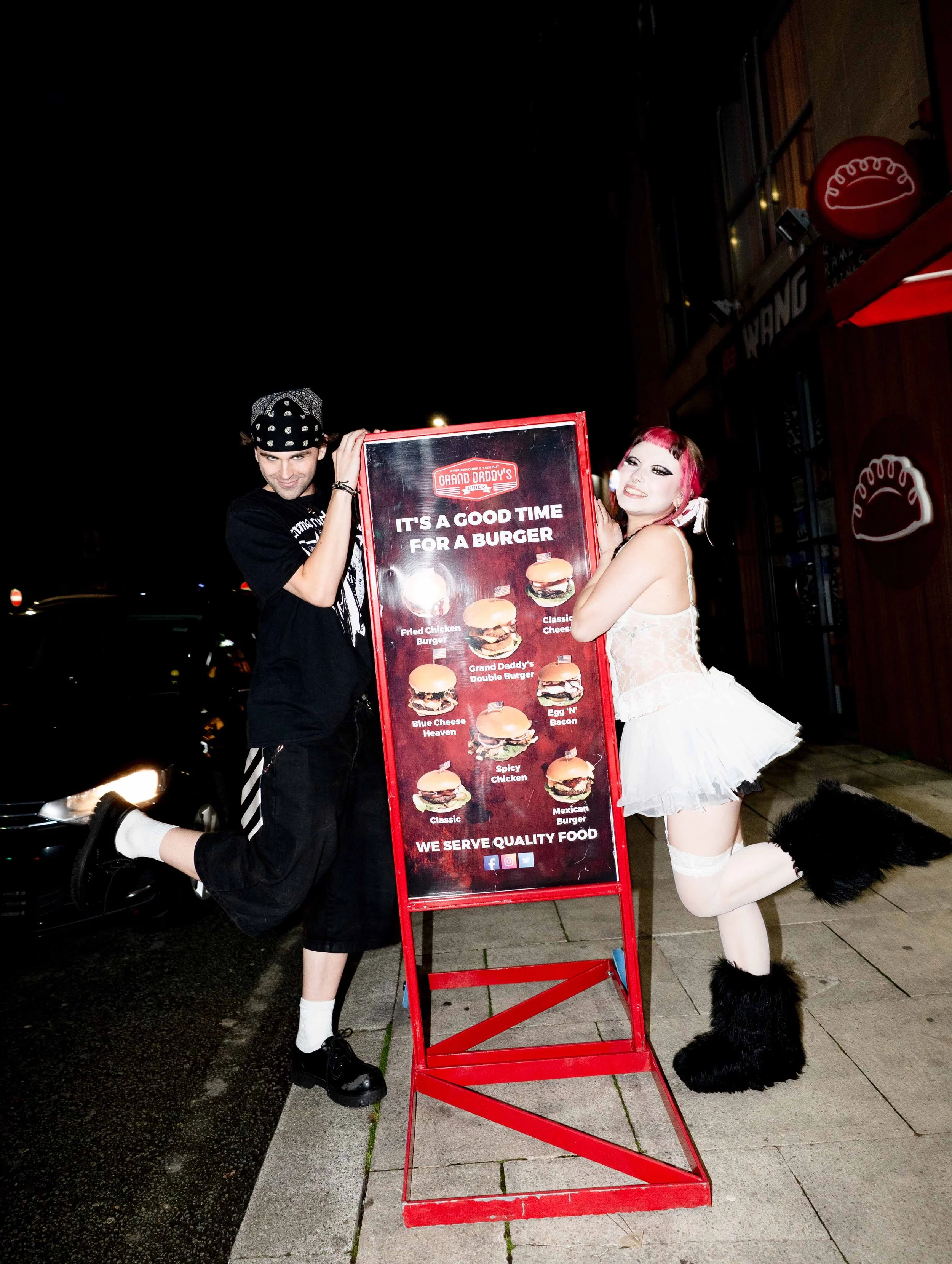 Two people, a man and a woman in a costume, standing on sidewalk at night beside a red sandwich board sign advertising burgers outside a restaurant.