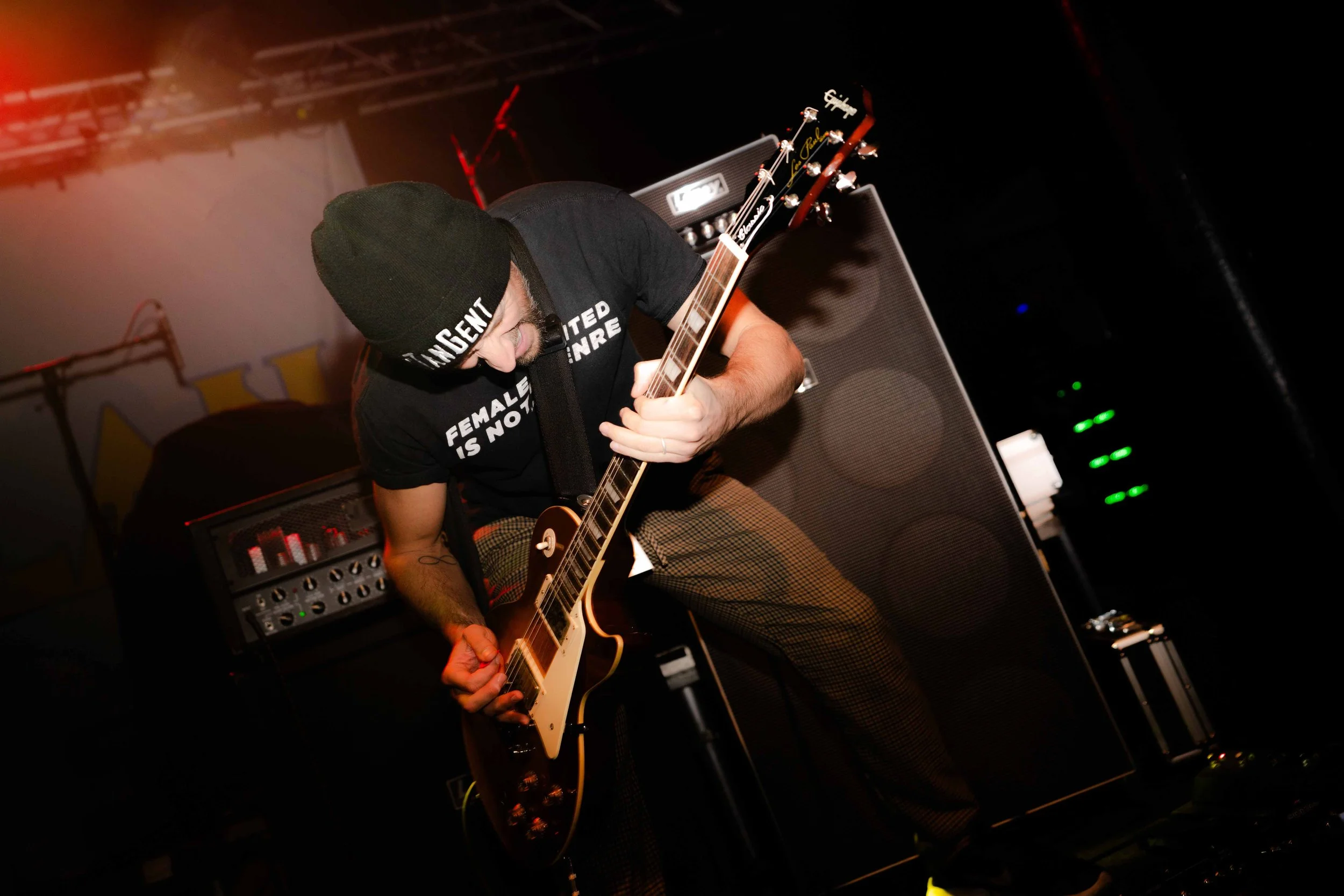 A musician wearing a black beanie and a black t-shirt playing an electric guitar on stage, with equipment and dark background lighting.