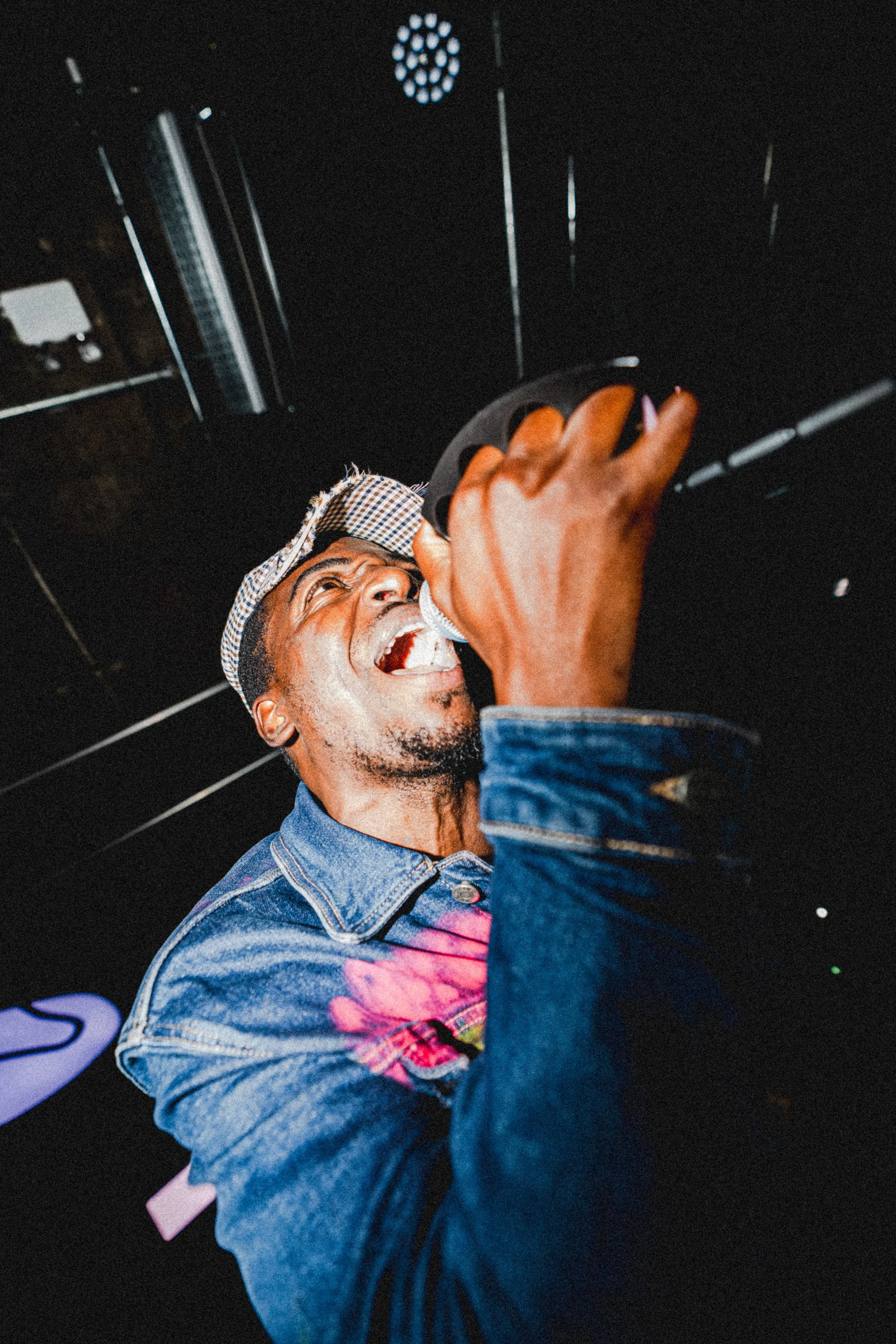 A man singing passionately into a microphone, wearing a denim jacket and a checkered cap, with a dark background.
