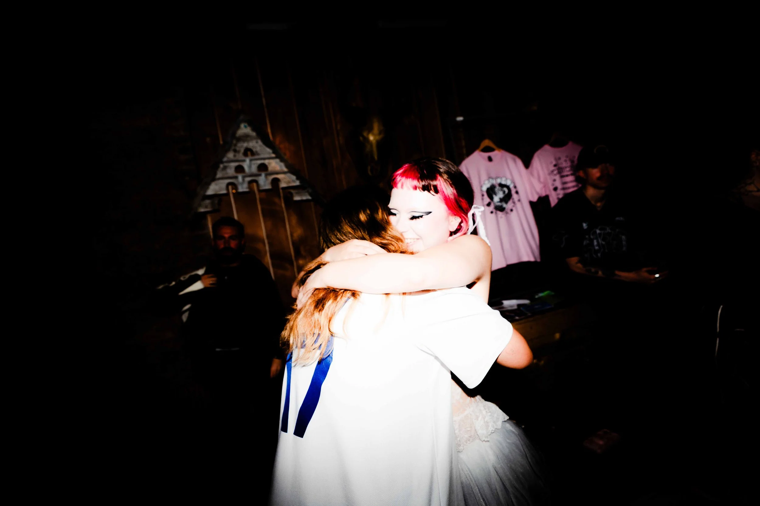 Two women embrace in a hug, smiling happily at an event with dark wood interior and pink T-shirts hanging in the background.
