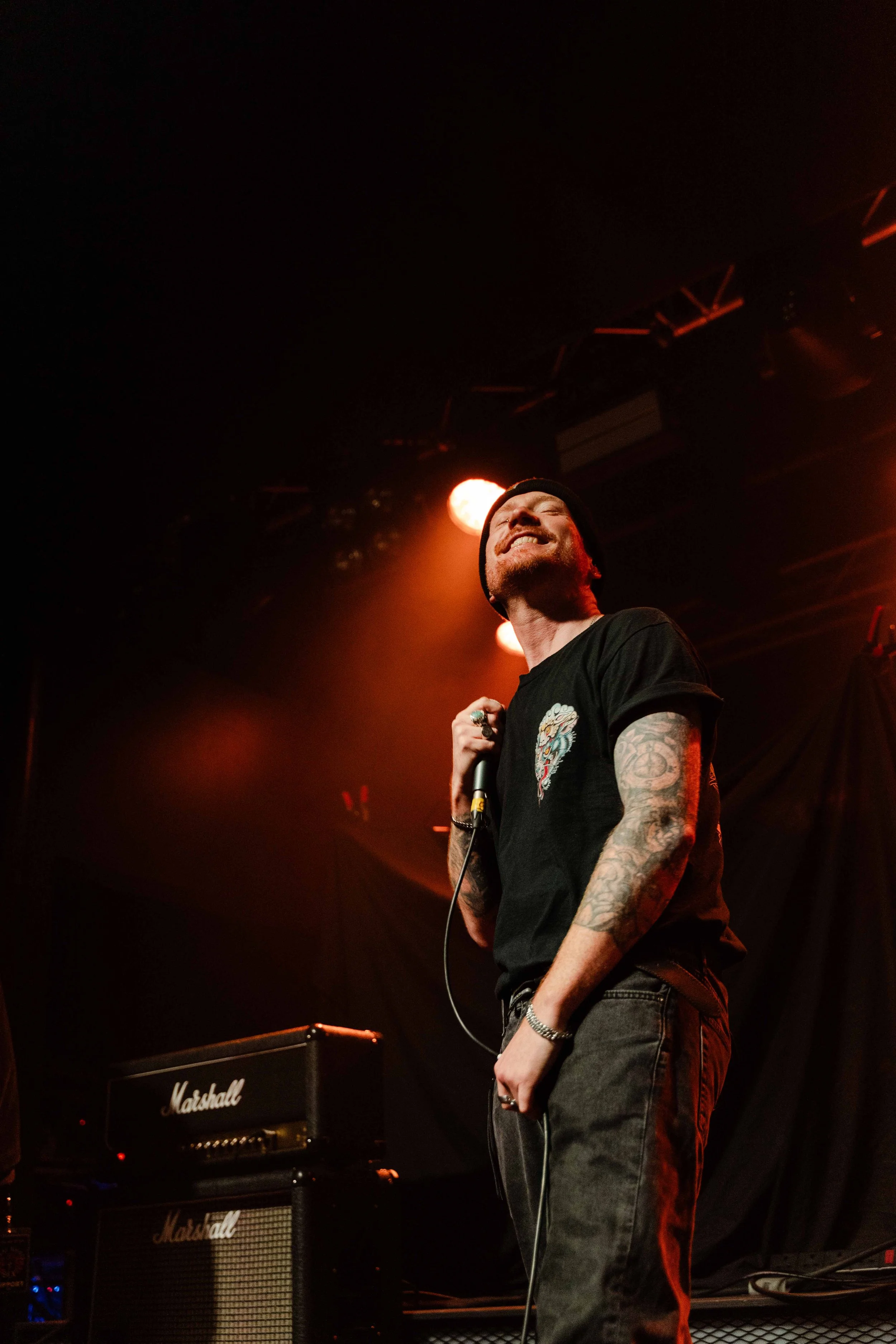 A man performing on stage, smiling, holding a microphone, with tattooed arms, wearing a black shirt, dark pants, and a beanie, with stage lights overhead.