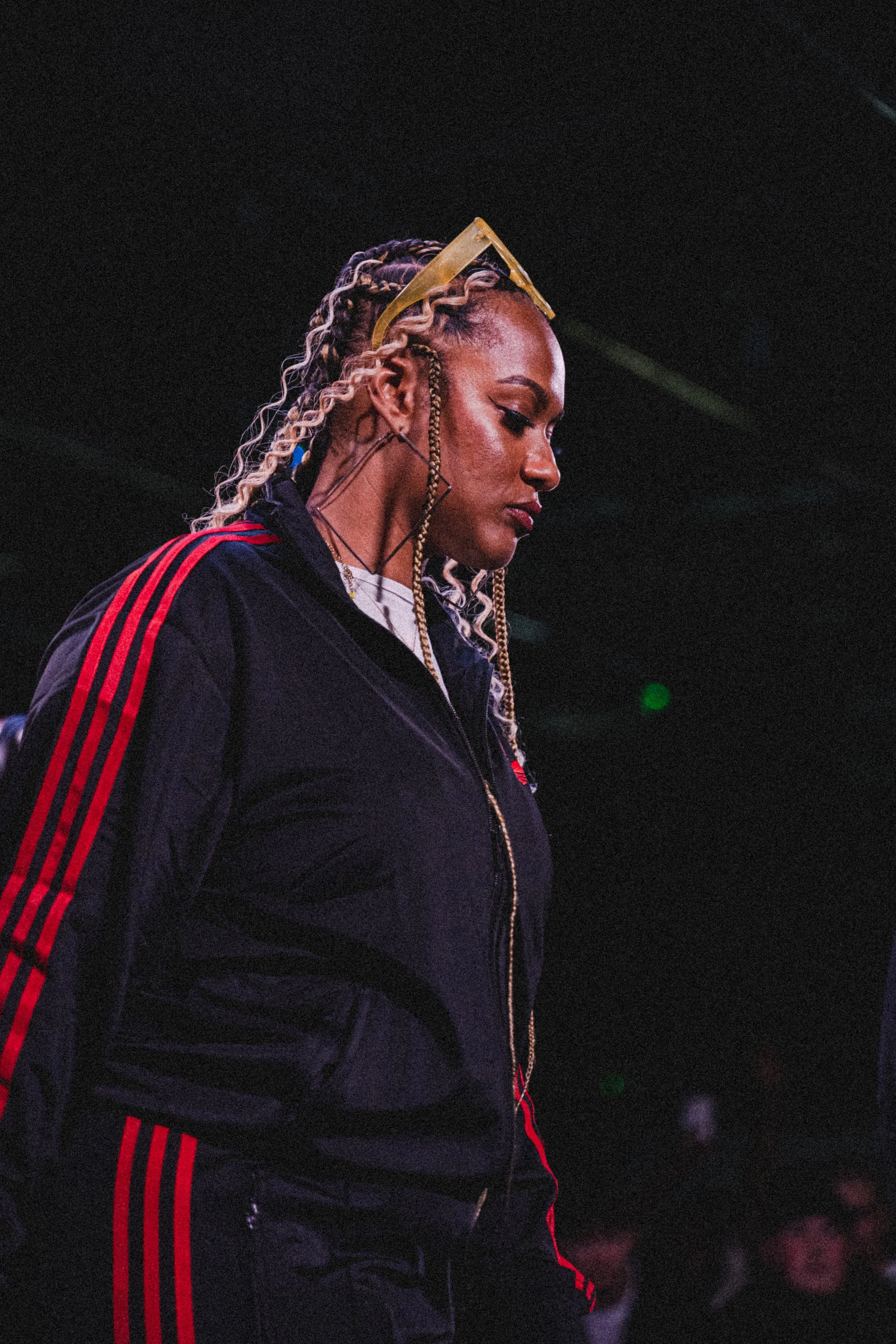 Female DJ wearing black and red Adidas tracksuit, with blonde braids, yellow sunglasses on head, performing at a dark venue.