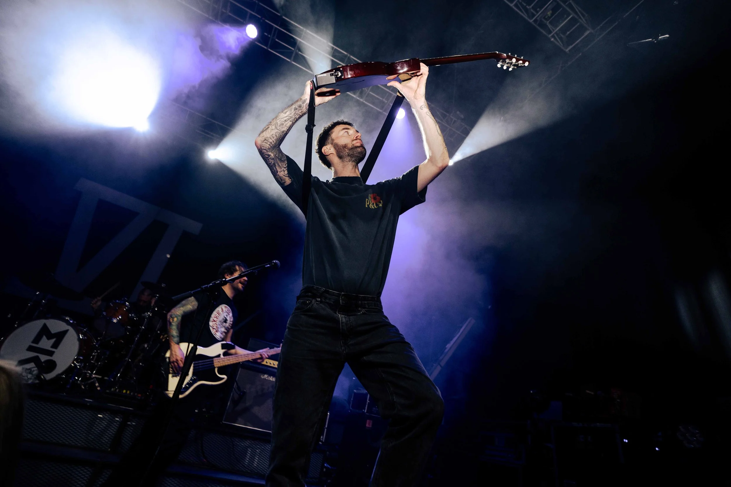 A musician on stage lifting a guitar above his head with both hands, while a drummer and another guitarist are in the background, surrounded by stage lights and smoke.