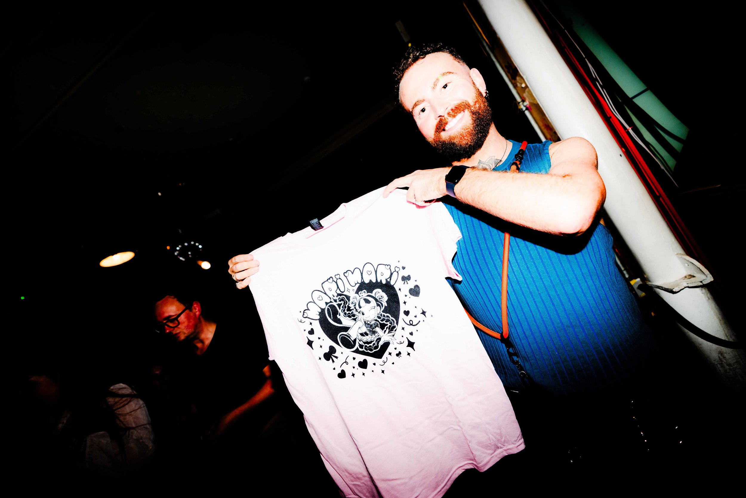 A smiling man with a beard holding a white T-shirt with an illustration of a girl and hearts, in a dark room with another person in the background.