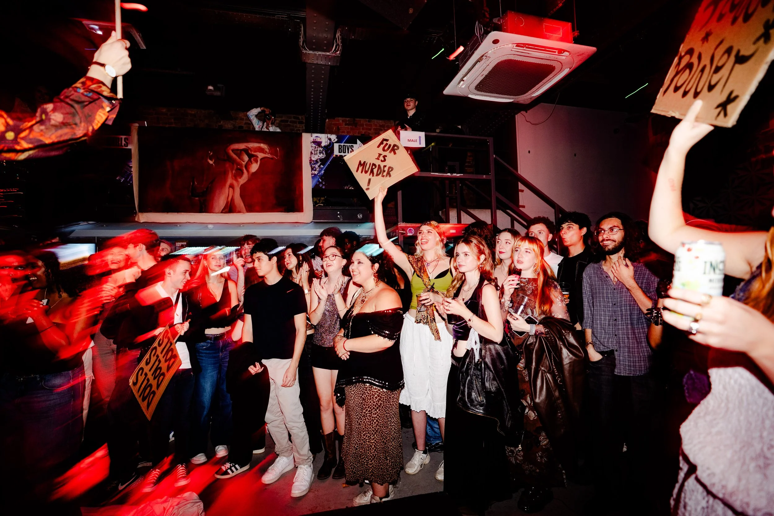 A group of young adults gathered indoors at a lively event, some holding signs like "Fur is Murder!" and "Power." The crowd is engaged and smiling, with some taking photos. The venue is dimly lit with red lighting, and there is a mezzanine area above