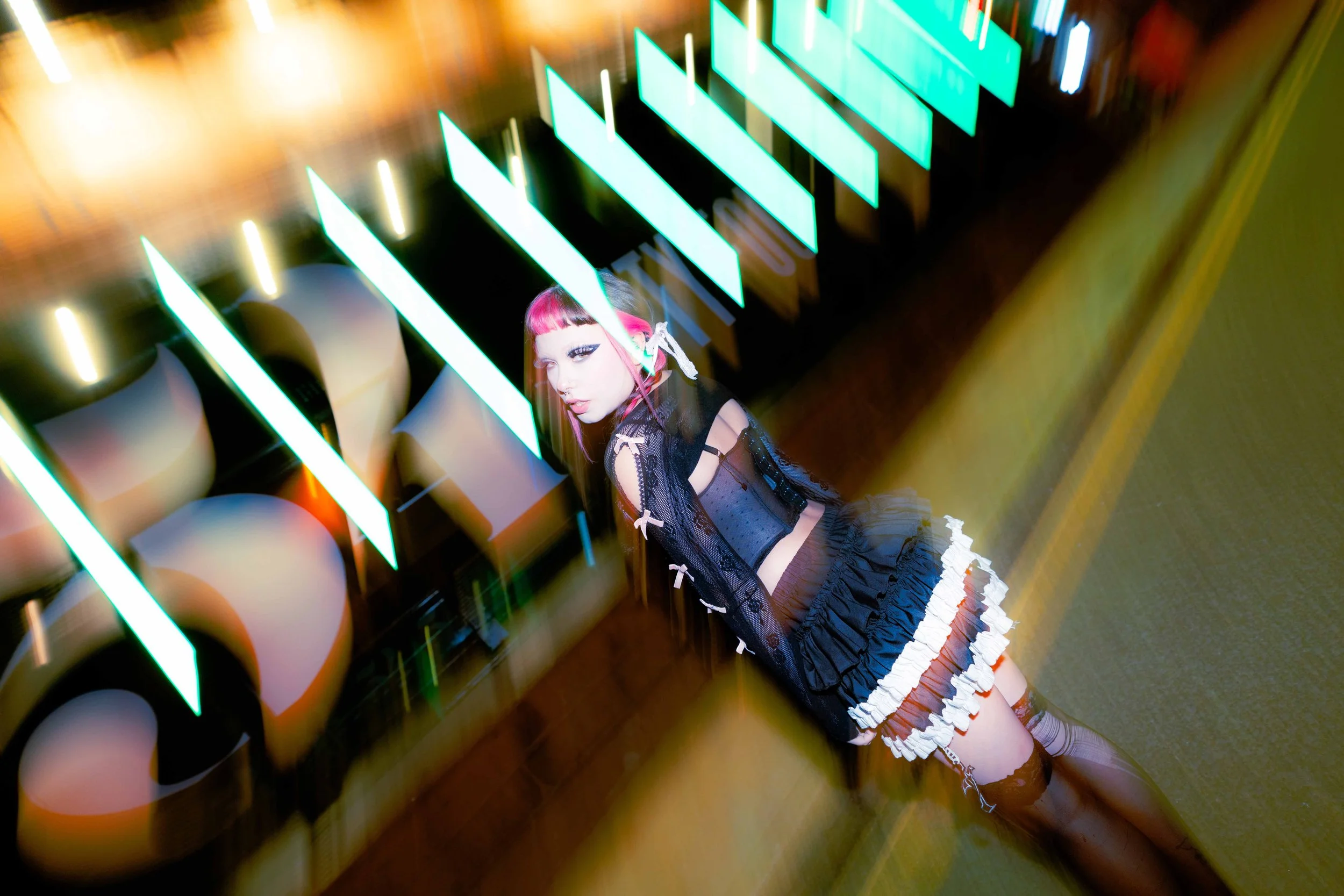 A woman with pink hair and gothic fashion style leaning against a neon-lit wall in a dimly lit room.
