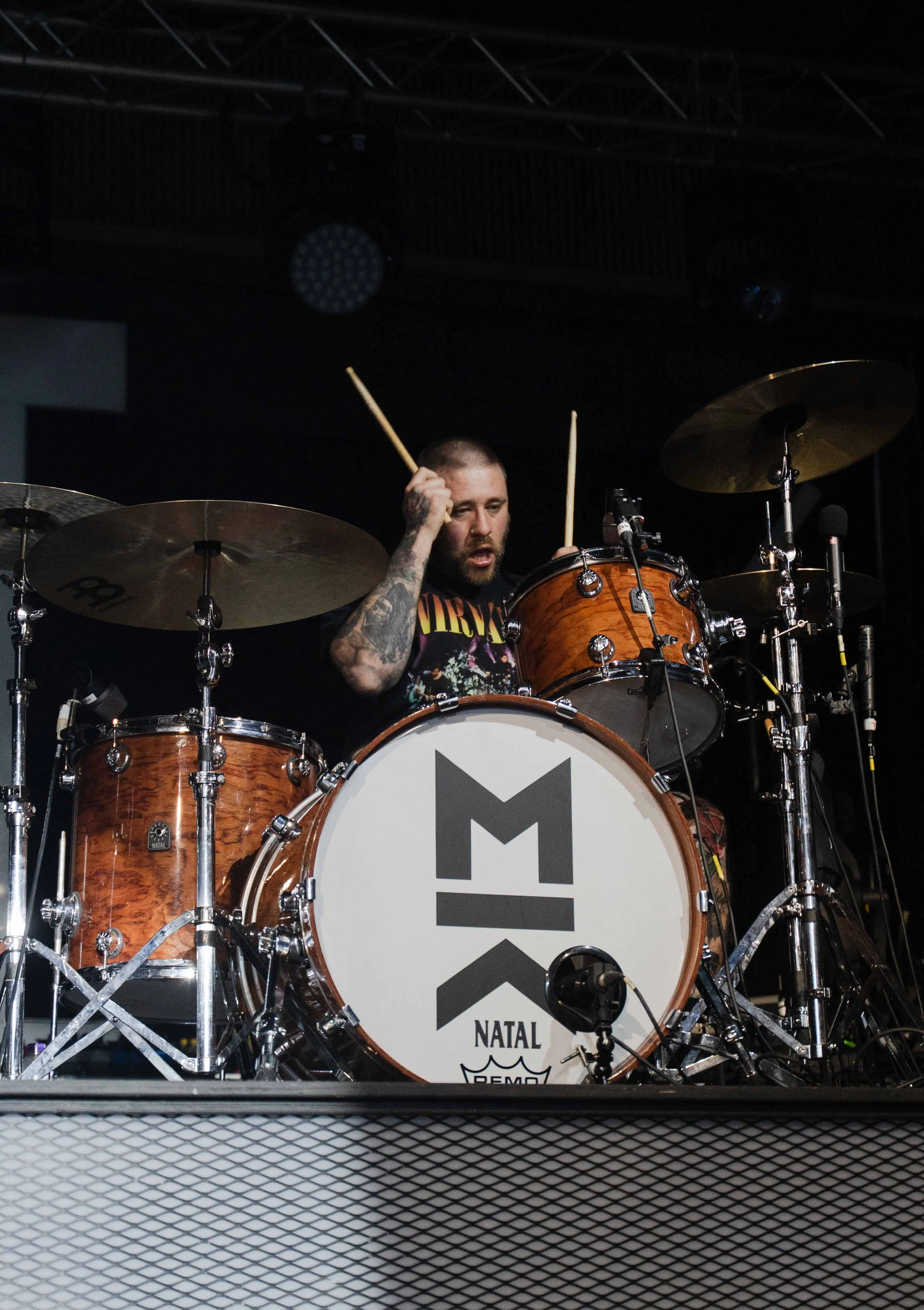 A man playing a drum set on stage, wearing a Nirvana T-shirt, with a bass drum that has the words "Natal" and "Remo" on it, and holding drumsticks in both hands.
