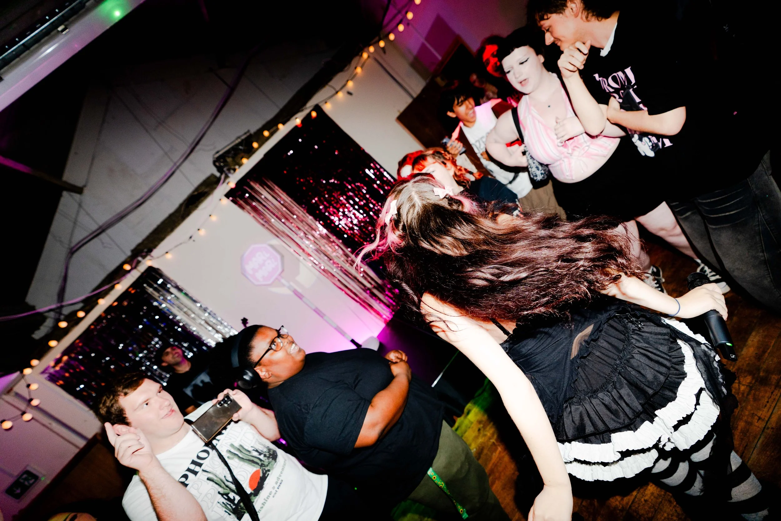 A group of people at a party, with a woman holding a microphone in the foreground, surrounded by friends dancing and socializing in a decorated room with string lights and shiny curtains.