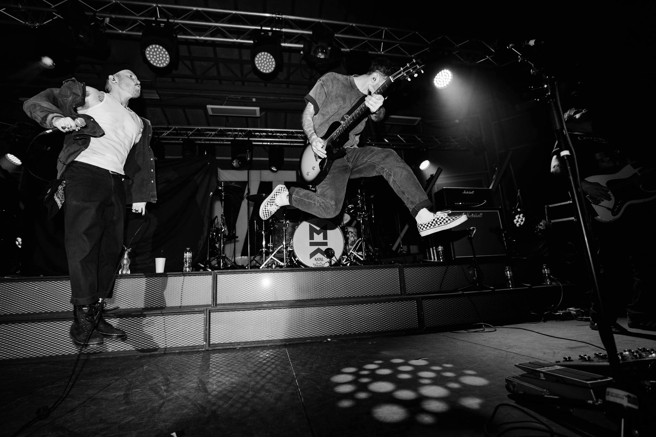 A black and white photo of a live music performance featuring a band on stage, with one member caught mid-air while jumping with a guitar, surrounded by a drum set, amplifiers, and other musicians.
