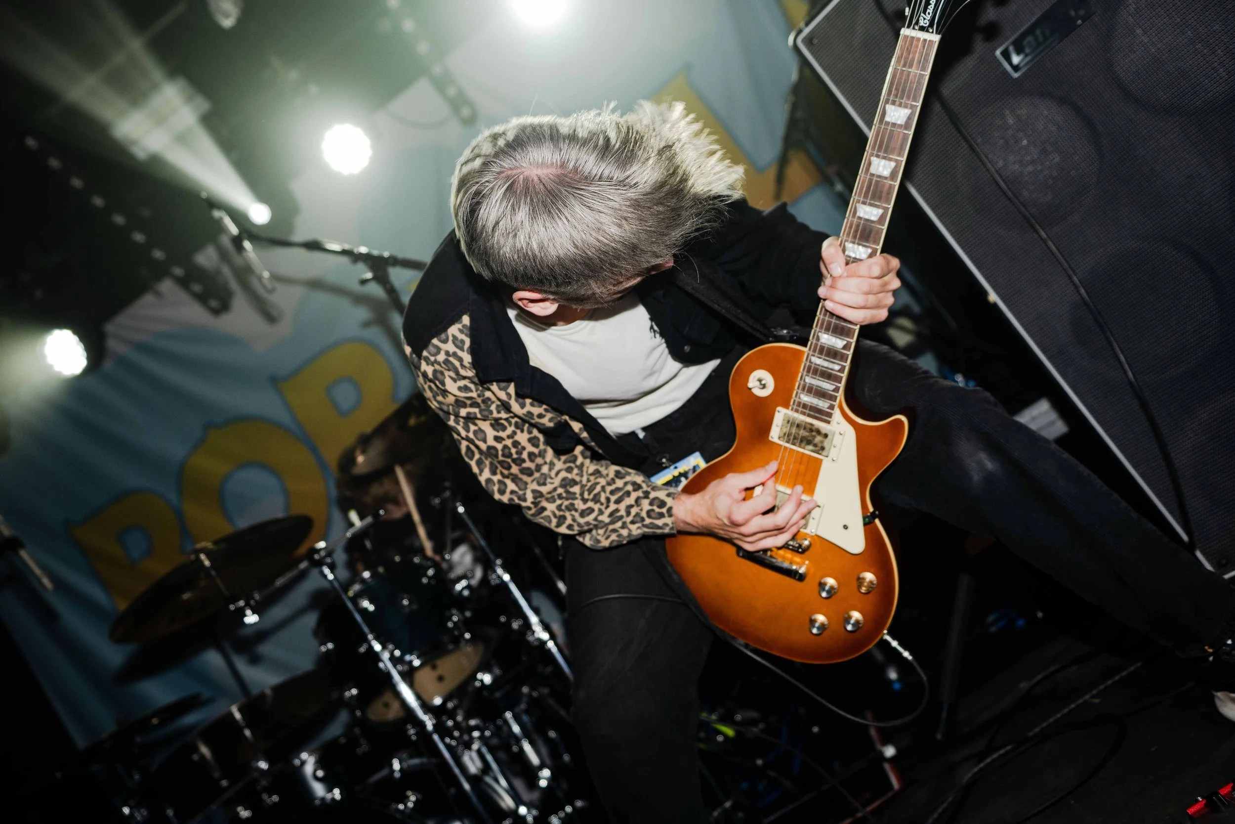 A musician with gray hair playing an orange electric guitar on stage, with a drum set and amplifiers in the background.