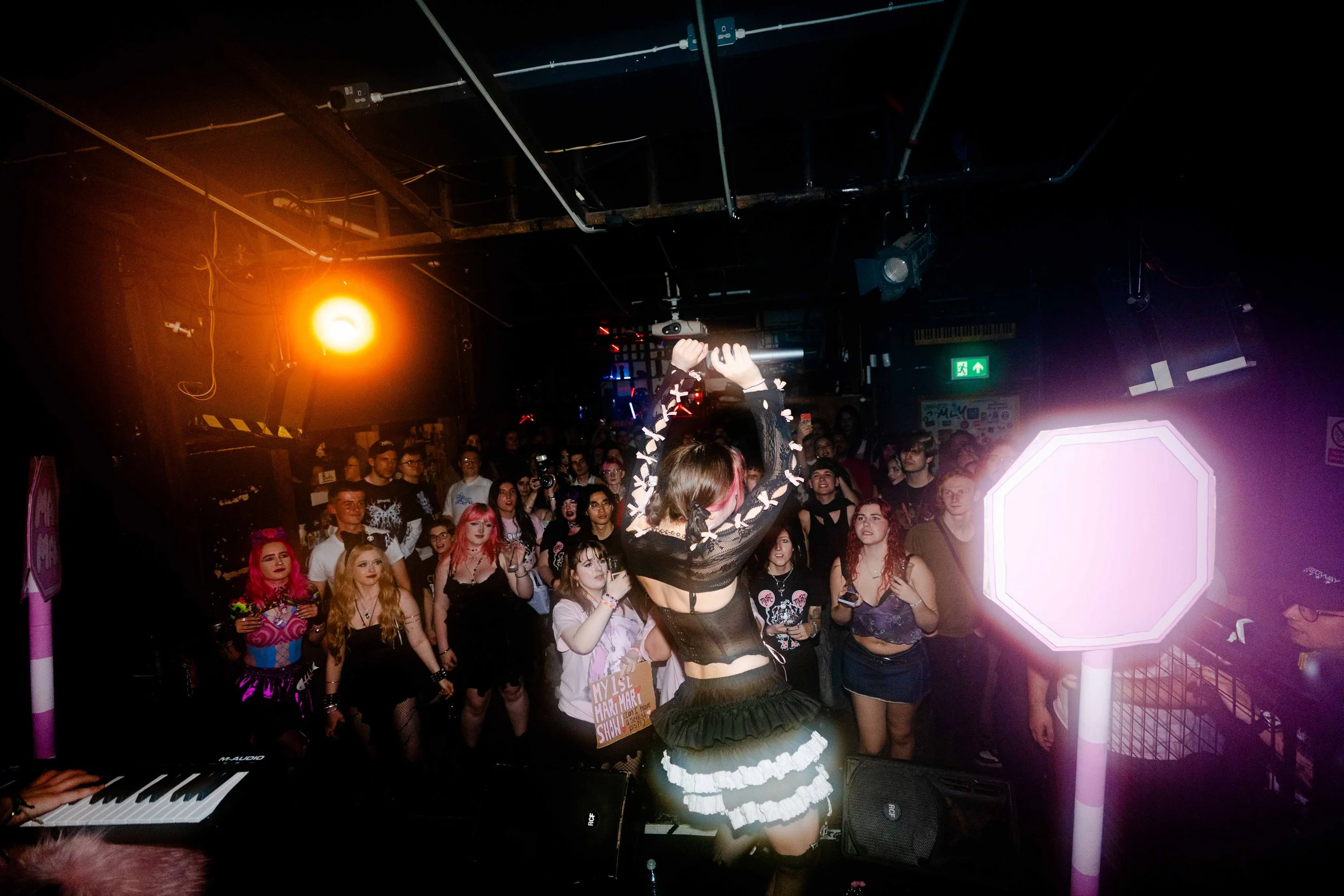 A performer dressed in black with white ruffles performs on stage with hands raised holding a microphone, while an audience watches at a concert or club with colorful lighting and a dark background.