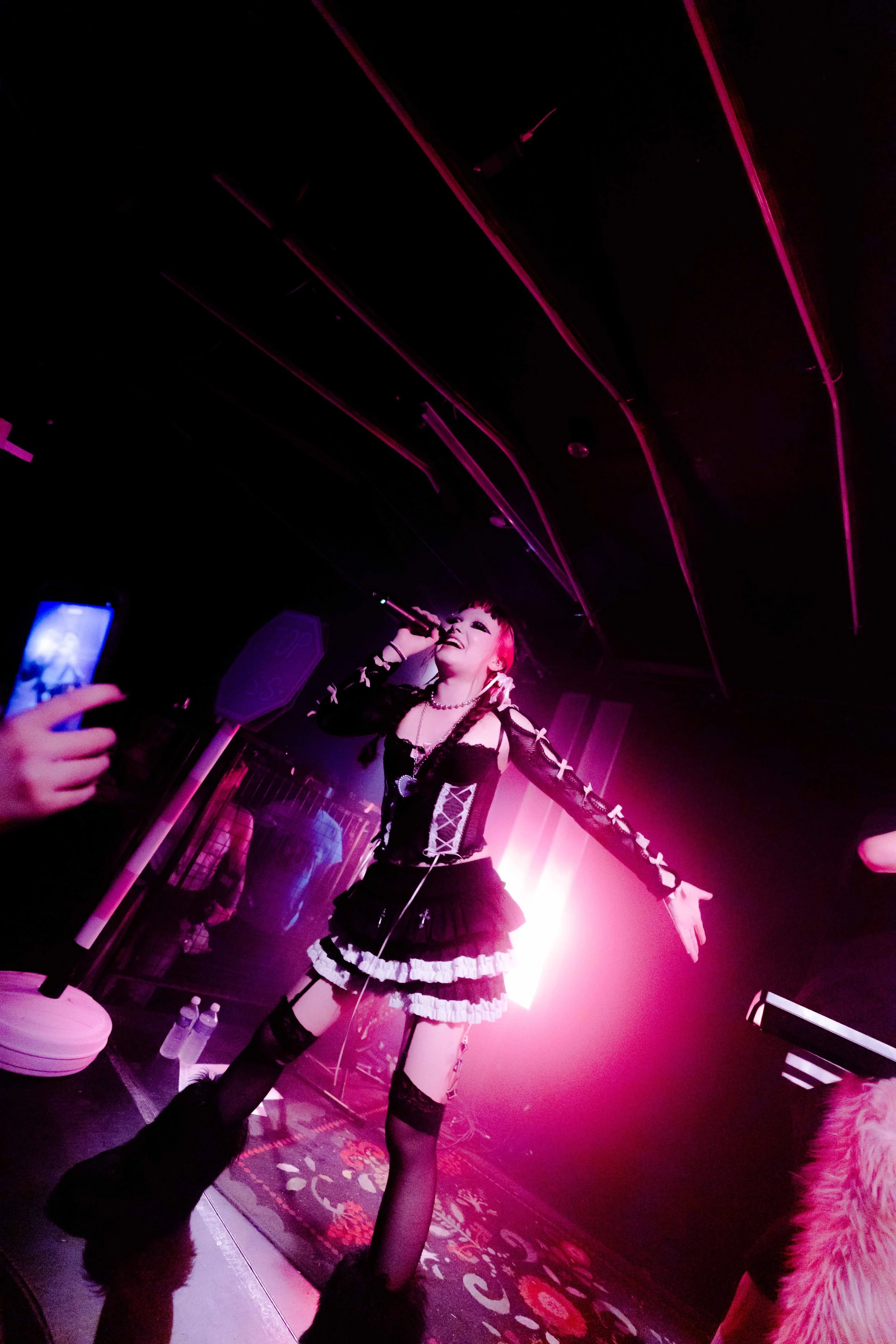 Performer singing into a microphone on stage with pink and purple lighting, wearing a goth-inspired black and white dress, stockings, and accessories. Audience members are taking pictures.