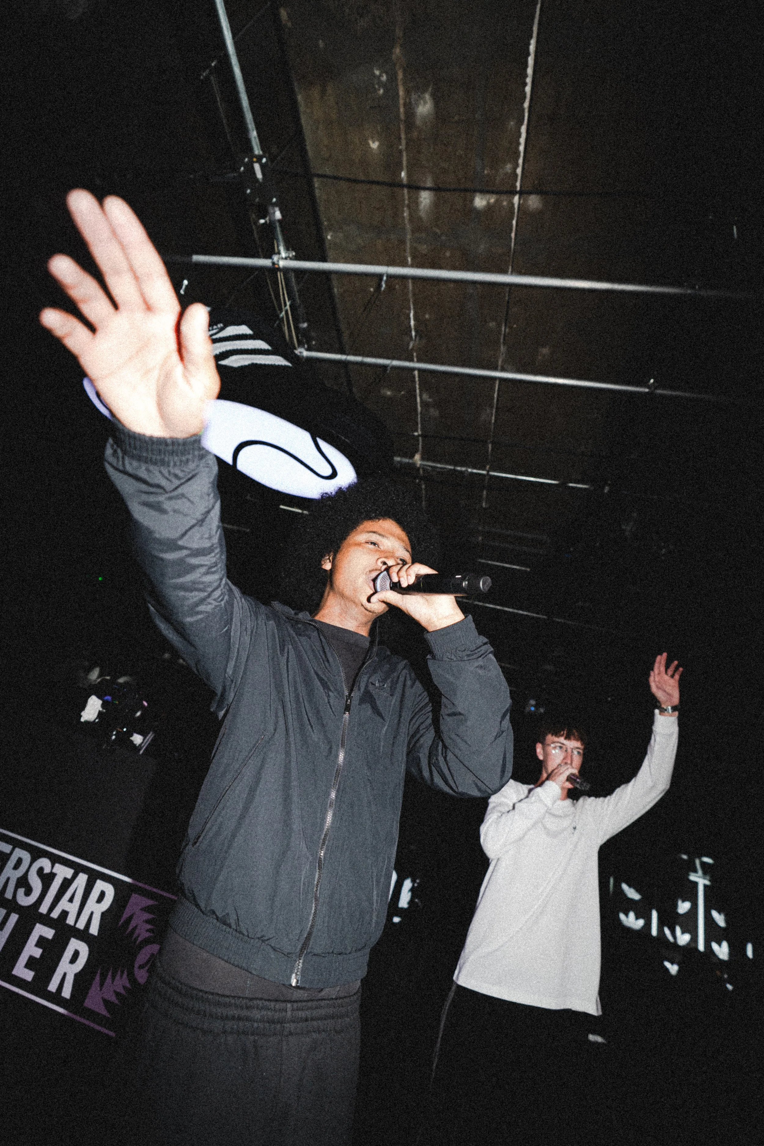 Two people singing into microphones, one in the foreground with his hand raised, on a stage with a dark background and purple signs that say 'Star' and 'Linger'.