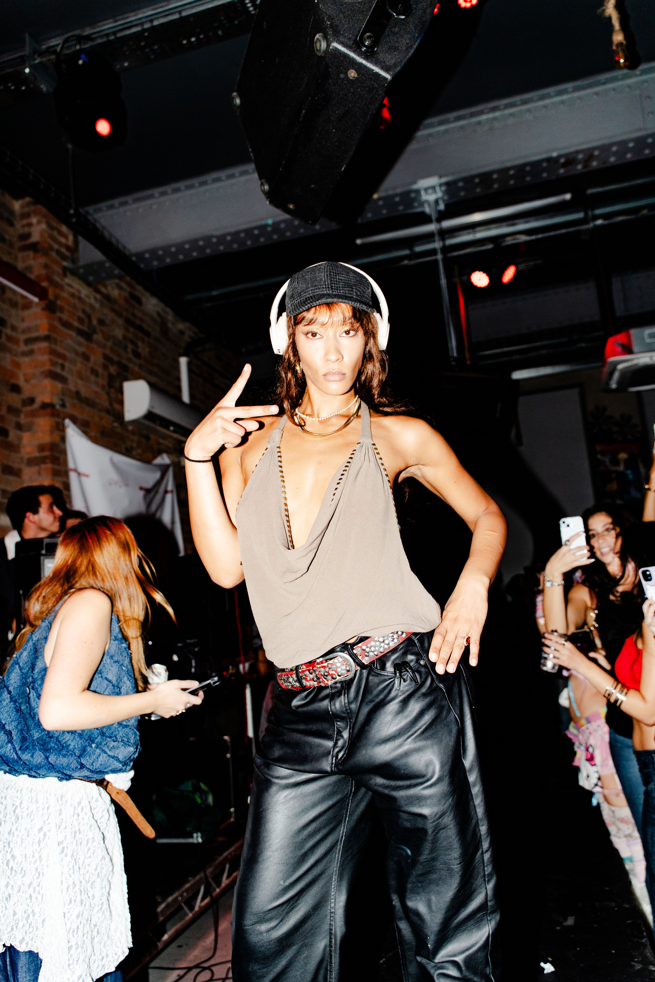 A woman in a cowl-neck top, black leather pants, and a baseball cap posing with a finger gun gesture at a crowded indoor event.