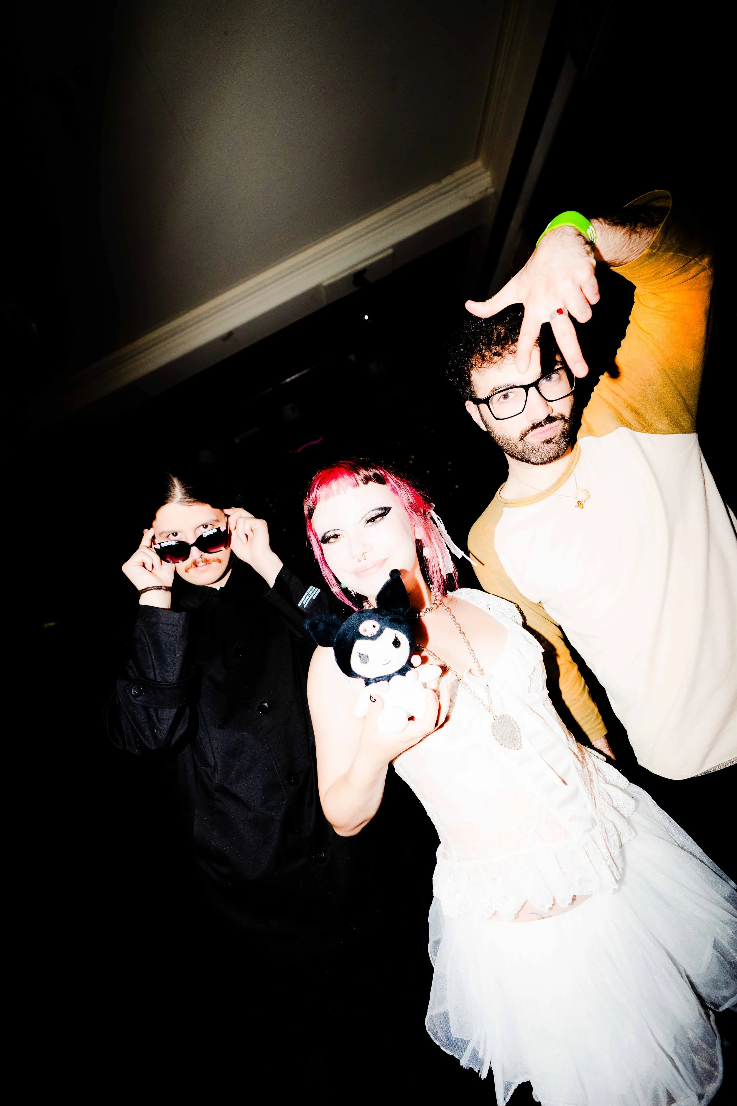 Three people posing in a dark room, one wearing sunglasses, another with pink hair holding a plush toy, and the third making a hand gesture near his face.