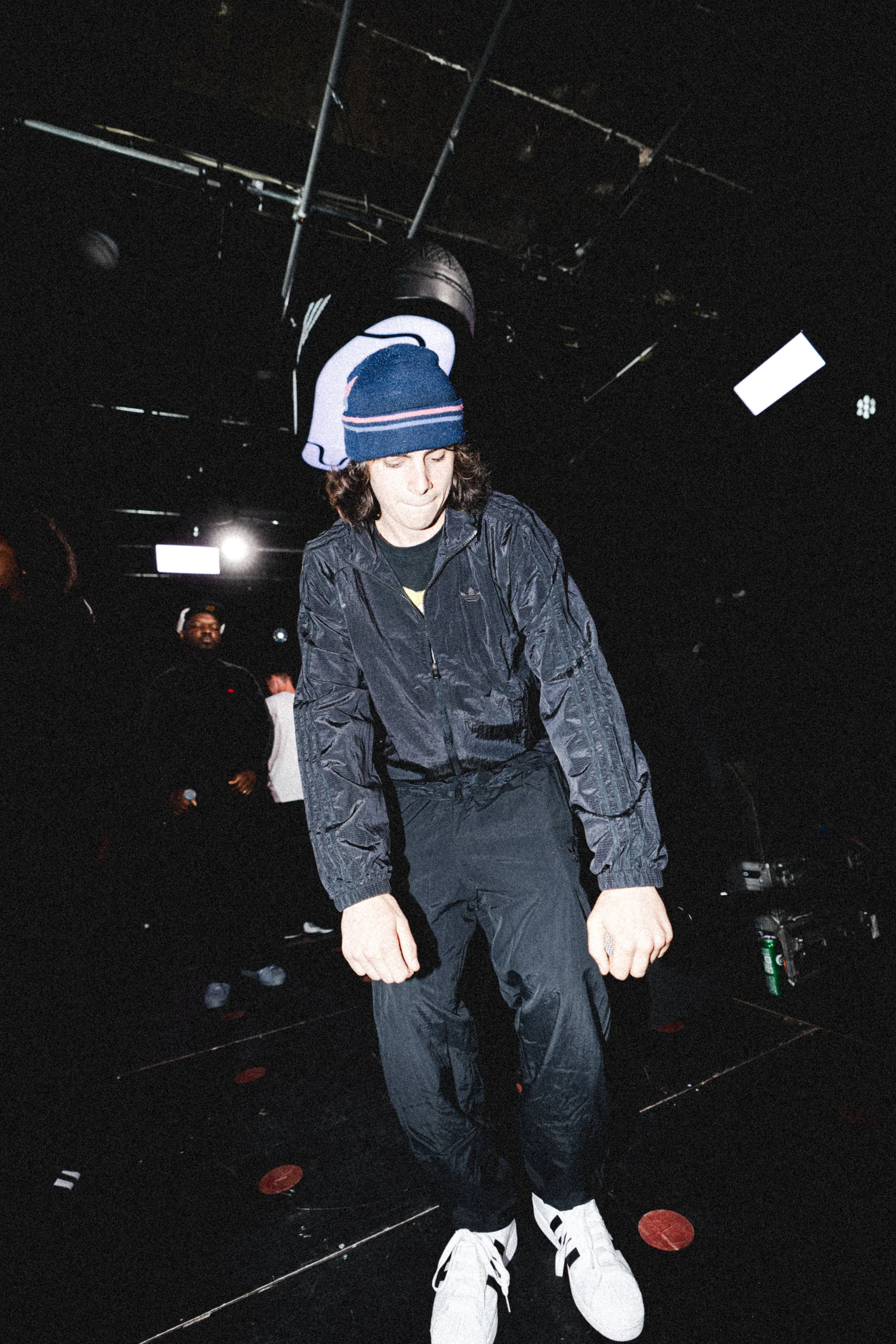 A young man wearing a blue knit cap, black jacket, and black pants is dancing or performing on stage with a black background. He is wearing white sneakers and appears to be in a club or concert setting.