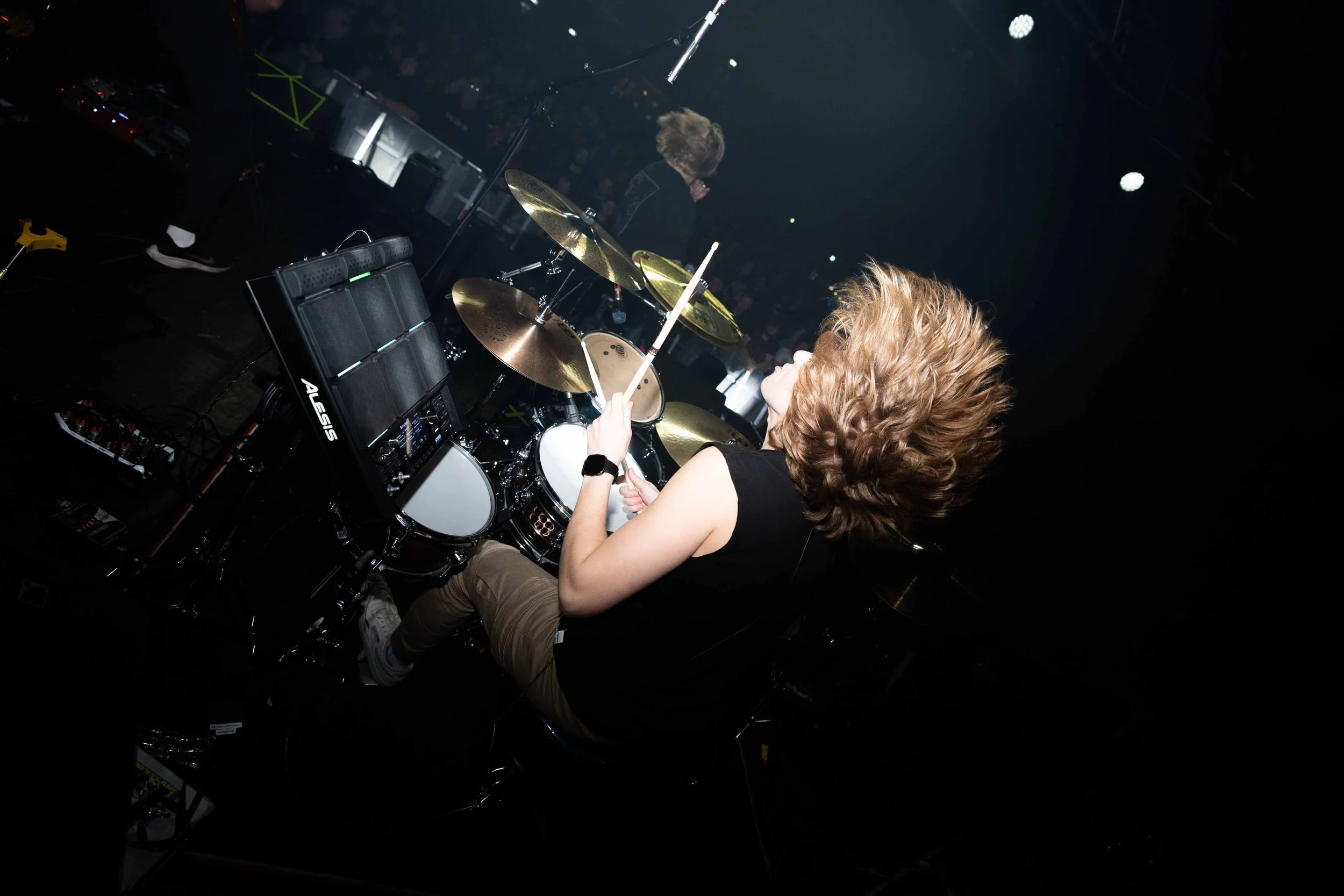 A female drummer with curly blonde hair playing a drum kit on stage during a concert, with a dark background and stage lights.