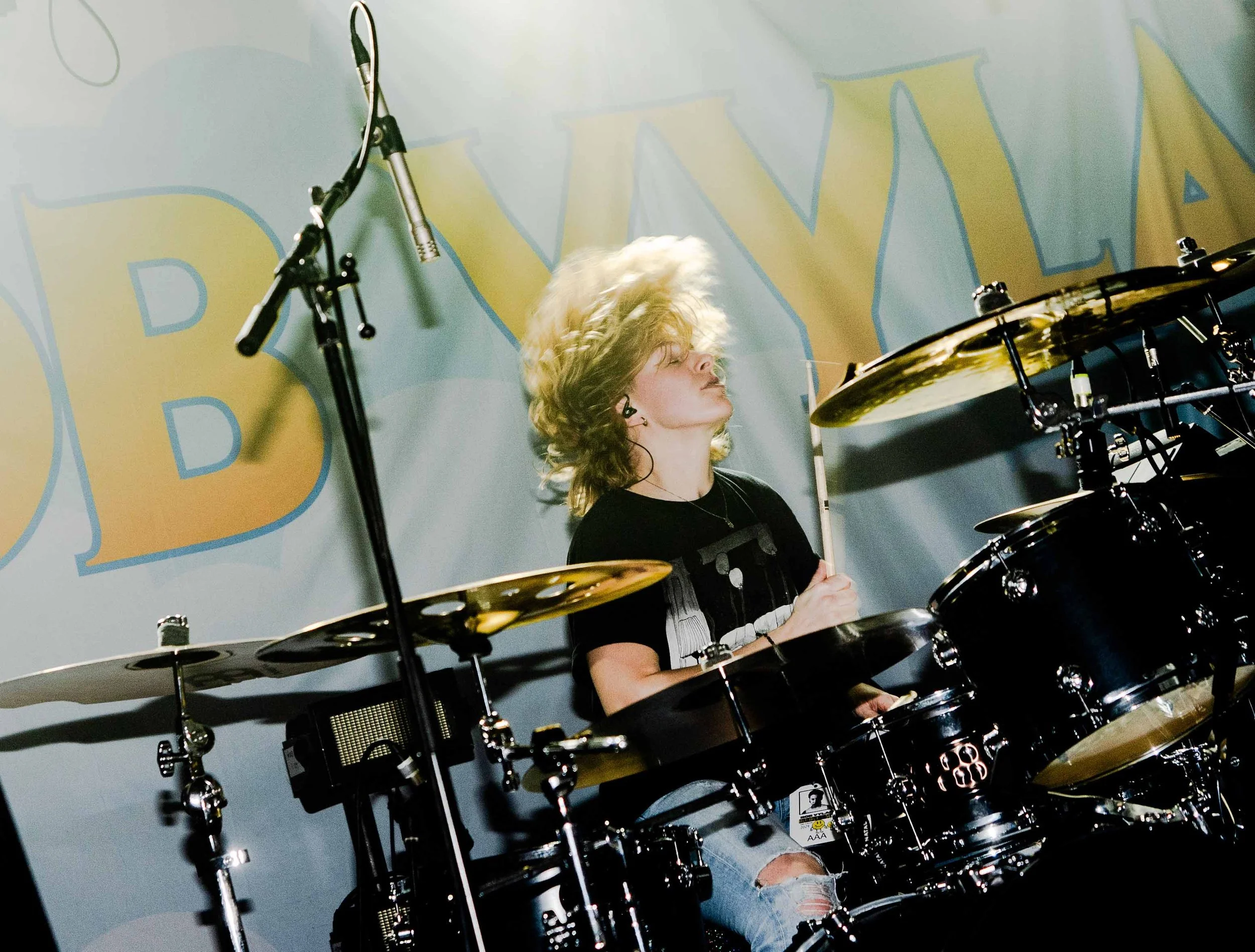 Young woman with curly blonde hair playing drums on stage, with large letters behind her.