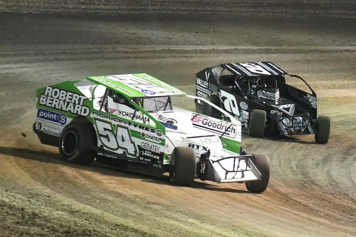 Two race cars on a dirt track, one white and green with the number 54, and the other black with the number 98, racing during a night event.
