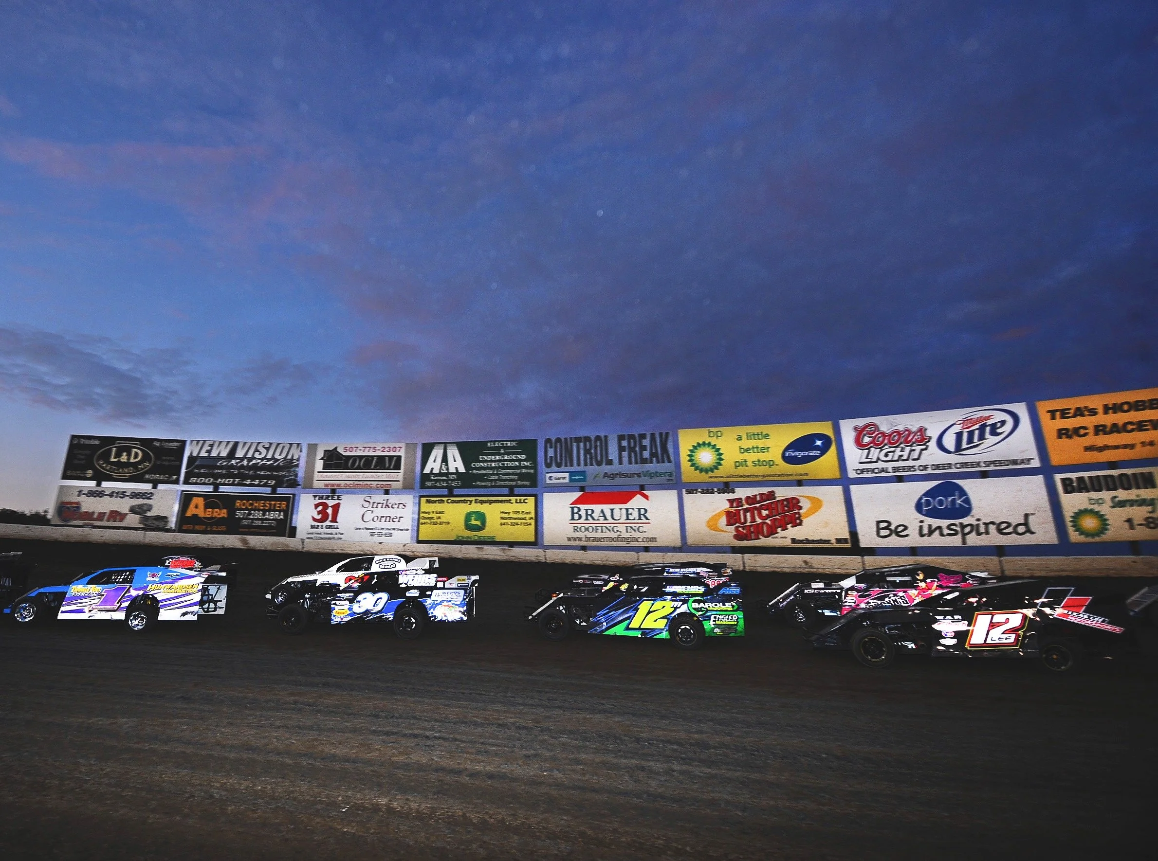 Daytime dirt track race with multiple race cars on the track, billboard advertisements in the background, and a partly cloudy sky overhead.