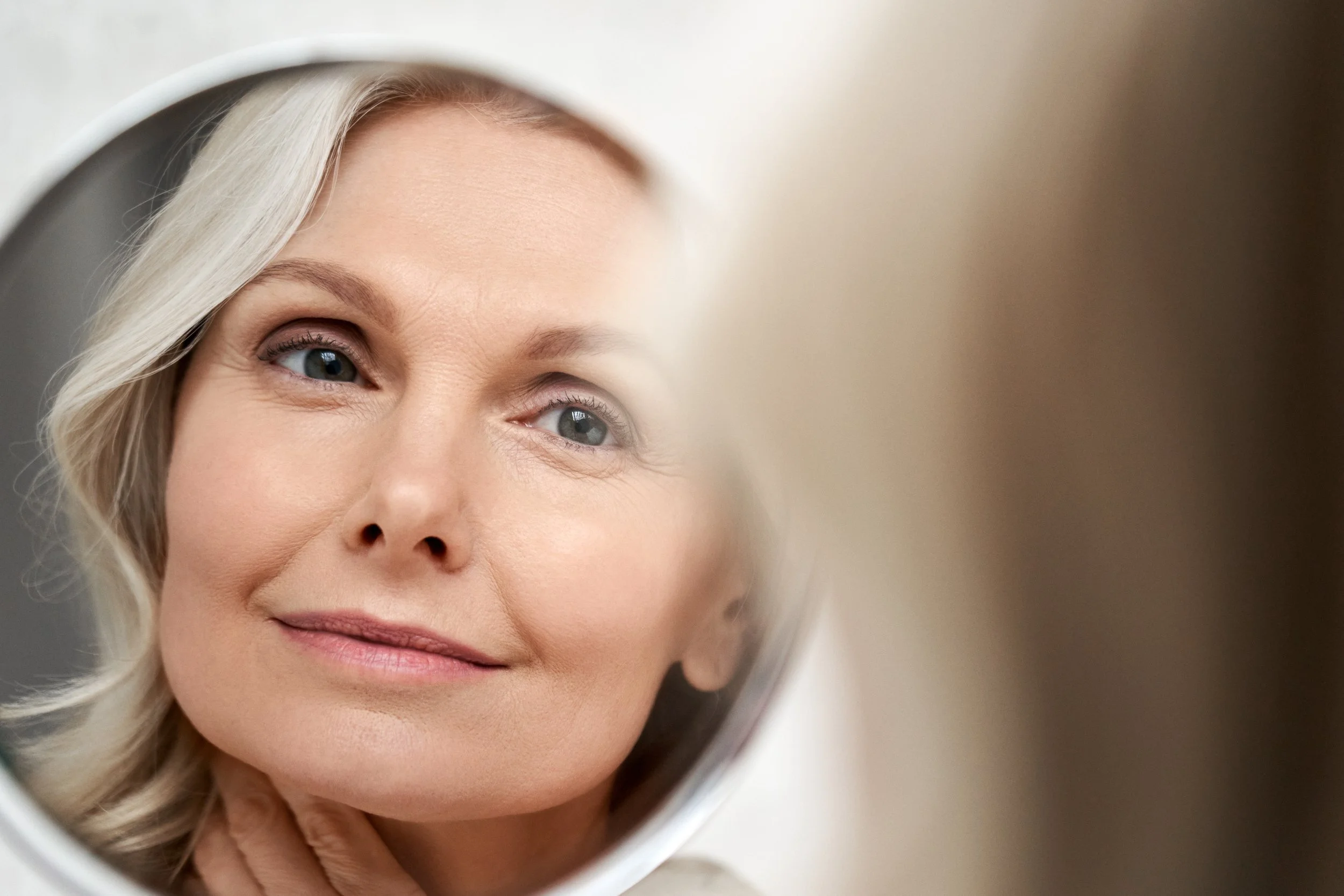 A woman with blonde hair looking into a mirror, with her reflection visible in the mirror.