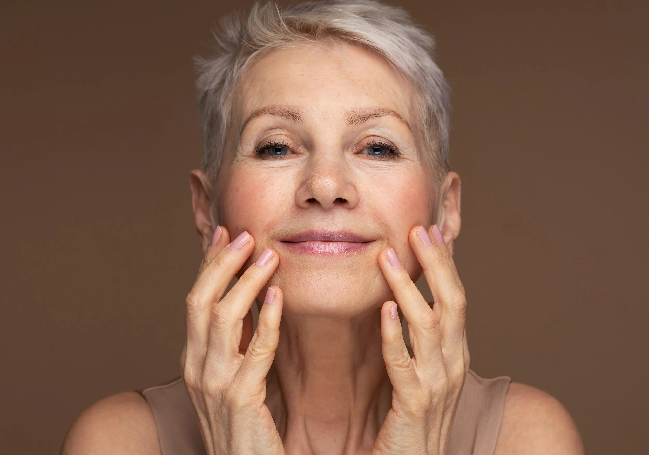 Close-up of a mature woman with short gray hair, smiling gently, touching her face with her fingers, against a neutral brown background.