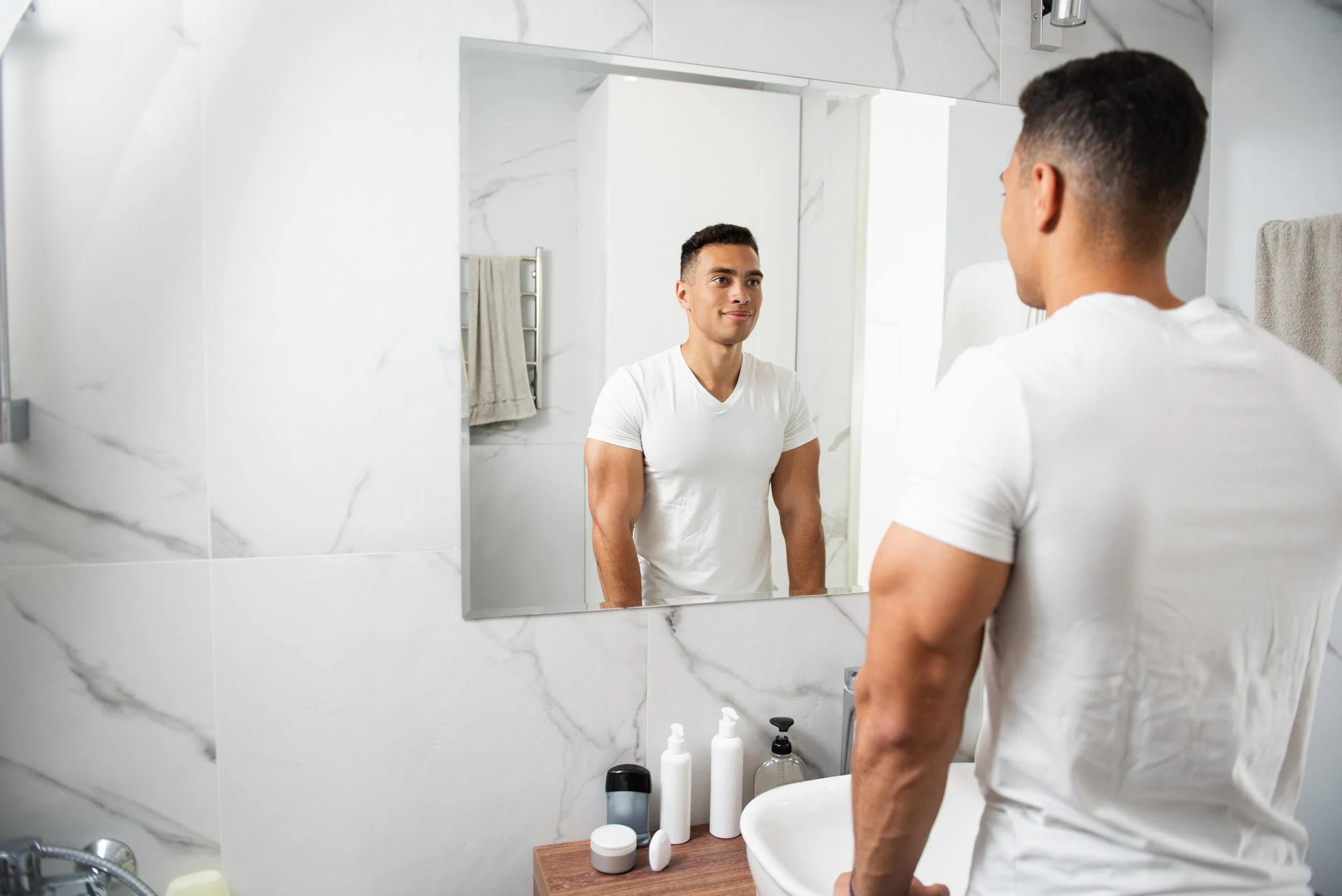 A young man stands in front of a bathroom mirror, looking at himself. The bathroom has white marble tiles, and there are various bottles and containers on a wooden countertop below the mirror.