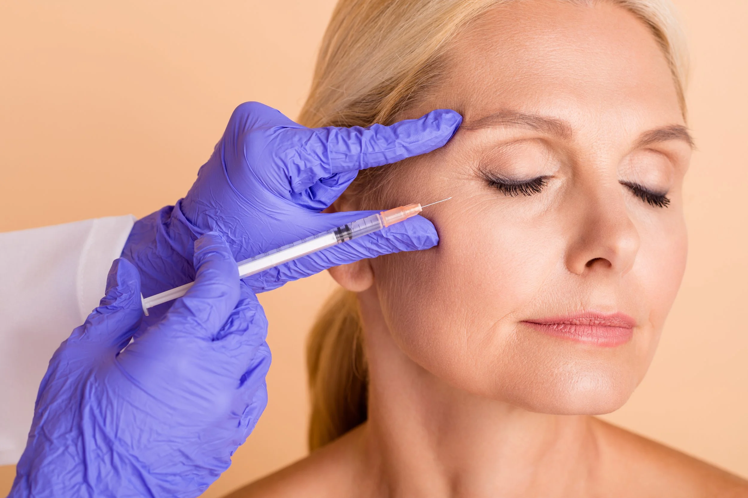 A healthcare professional wearing purple gloves administering a cosmetic or Botox injection into a woman's forehead.
